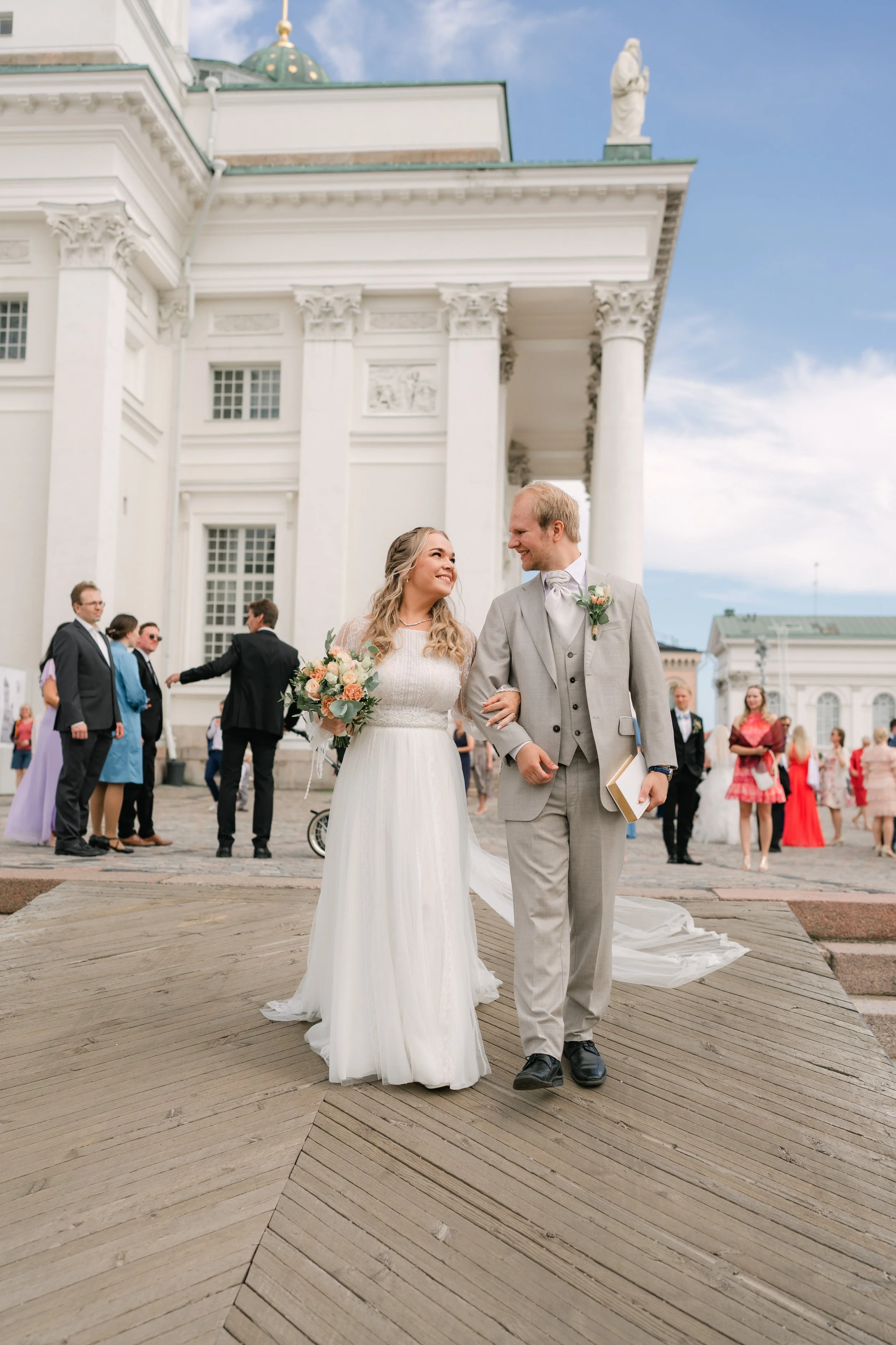 Bride and groom walking arm in arm outside a white classical building with wedding guests in the background.