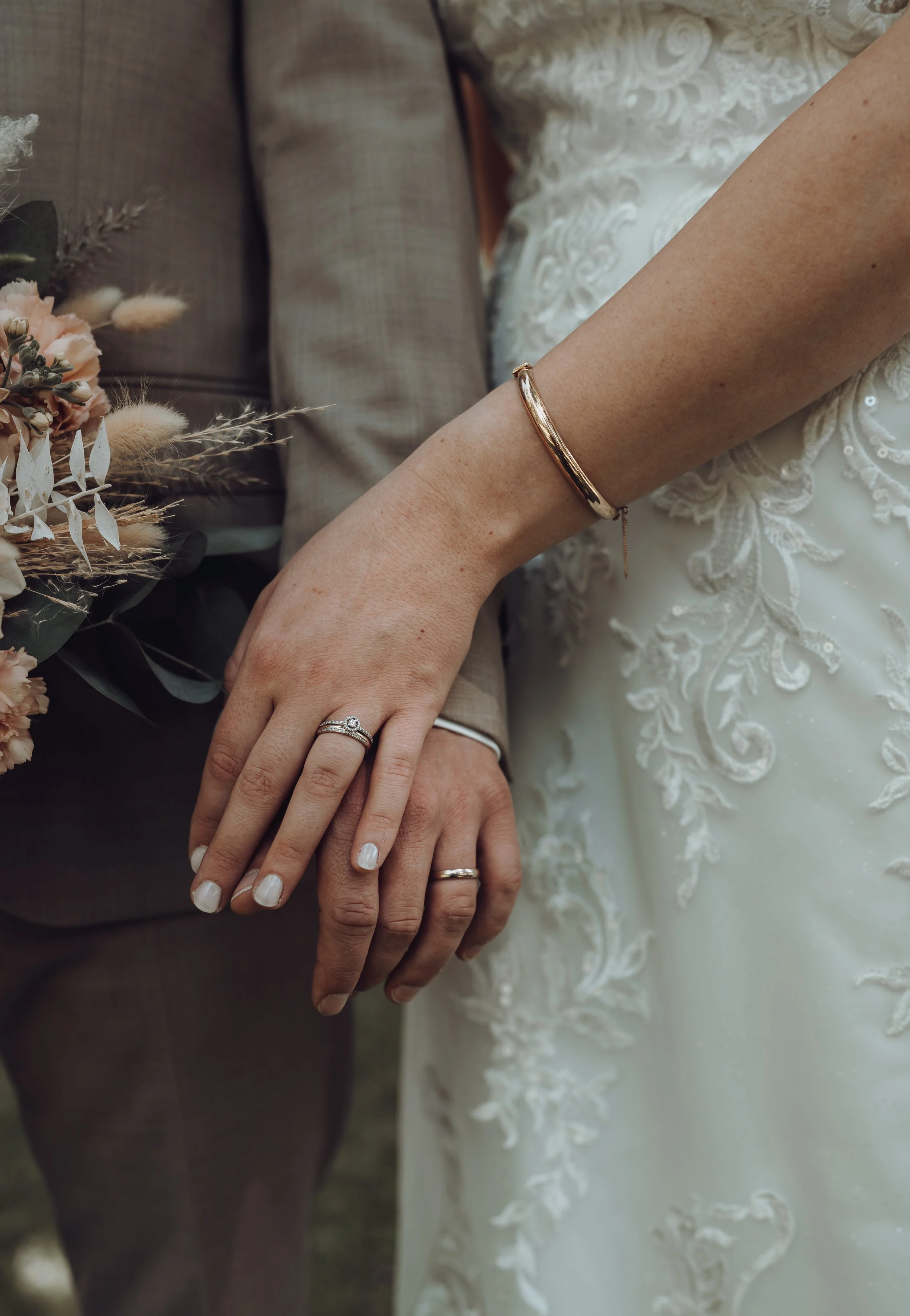 Close-up of bride and groom holding hands, wearing wedding rings, with lace dress and flower bouquet