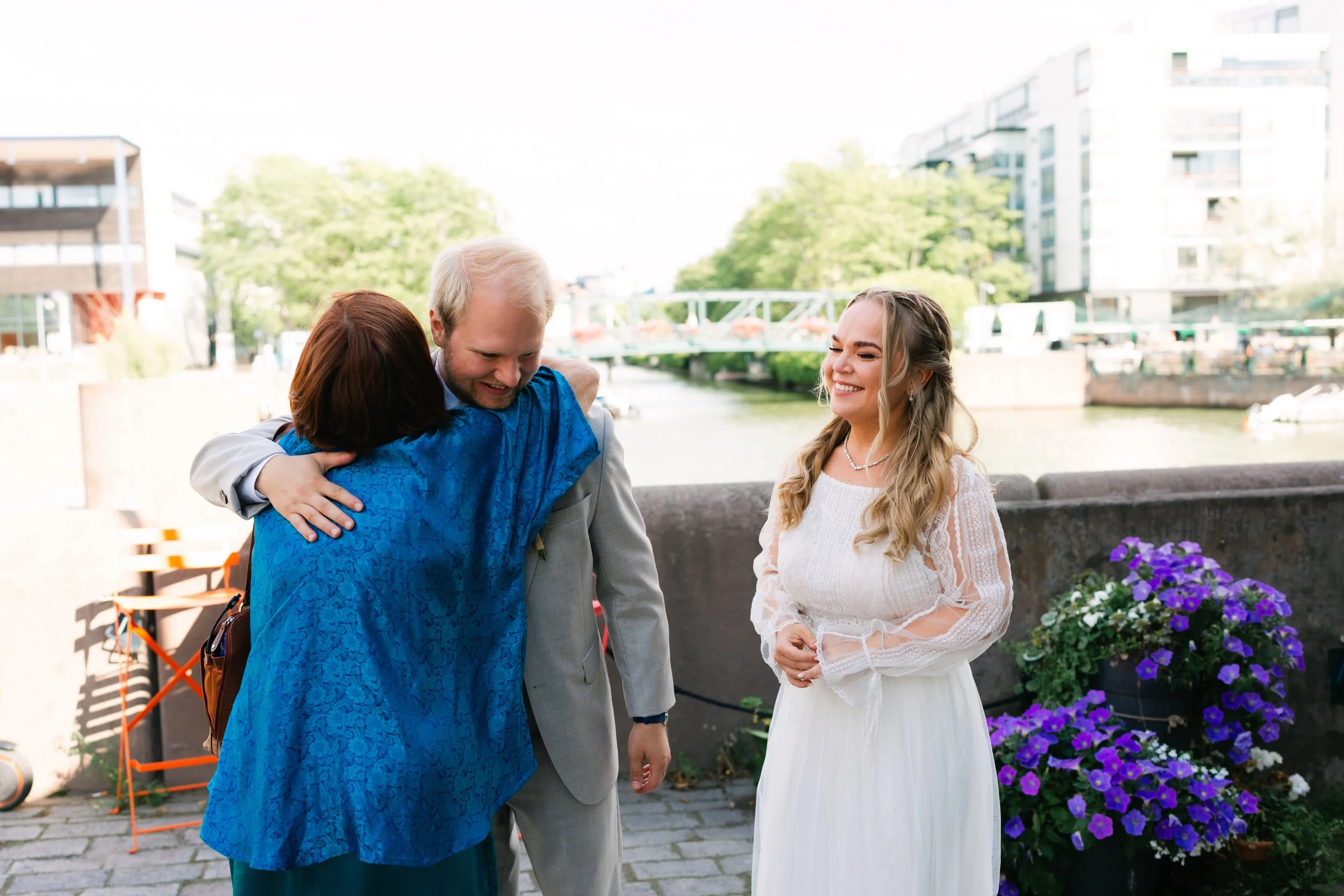 A woman hugging a man in a suit, with a smiling bride in a white dress nearby. The scene is outdoors by a river with purple flowers and modern buildings in the background.