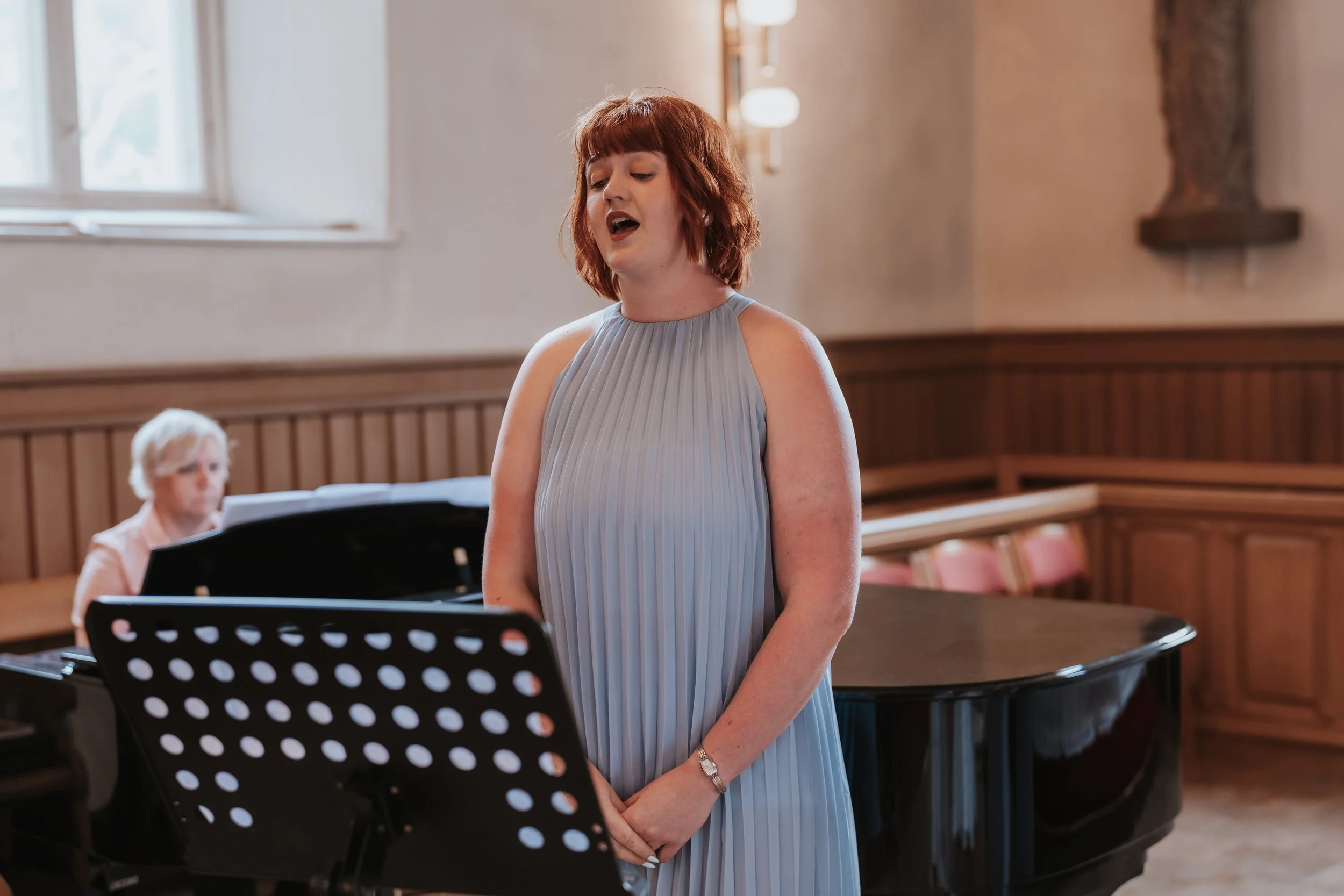 Woman singing with sheet music on stand, accompanied by pianist in room with large window.