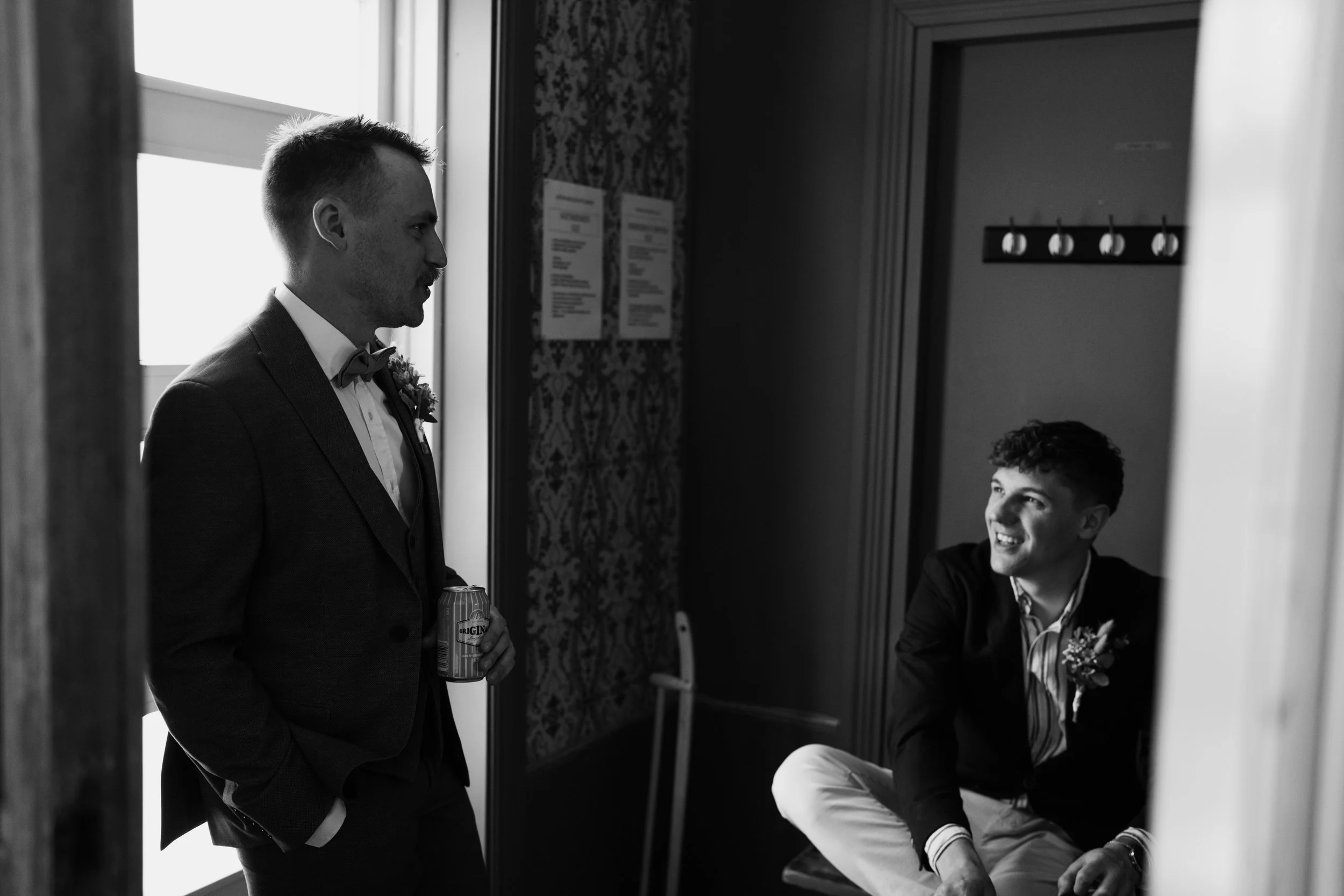 Two men in suits having a conversation indoors, with one holding a drink, in a black and white photo. The atmosphere seems formal, possibly a wedding or event.