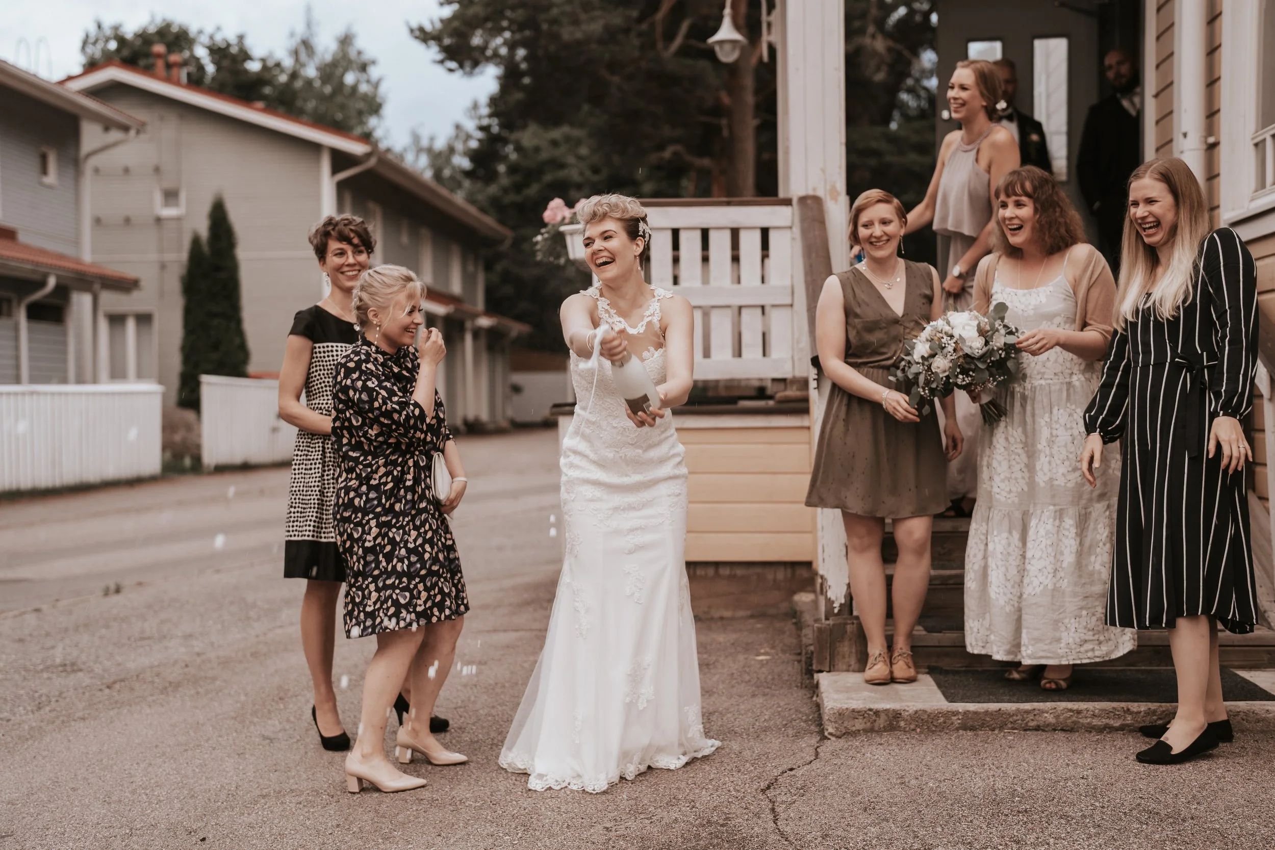 Bride in white dress popping champagne outside, surrounded by joyful people, including a woman holding a bouquet.