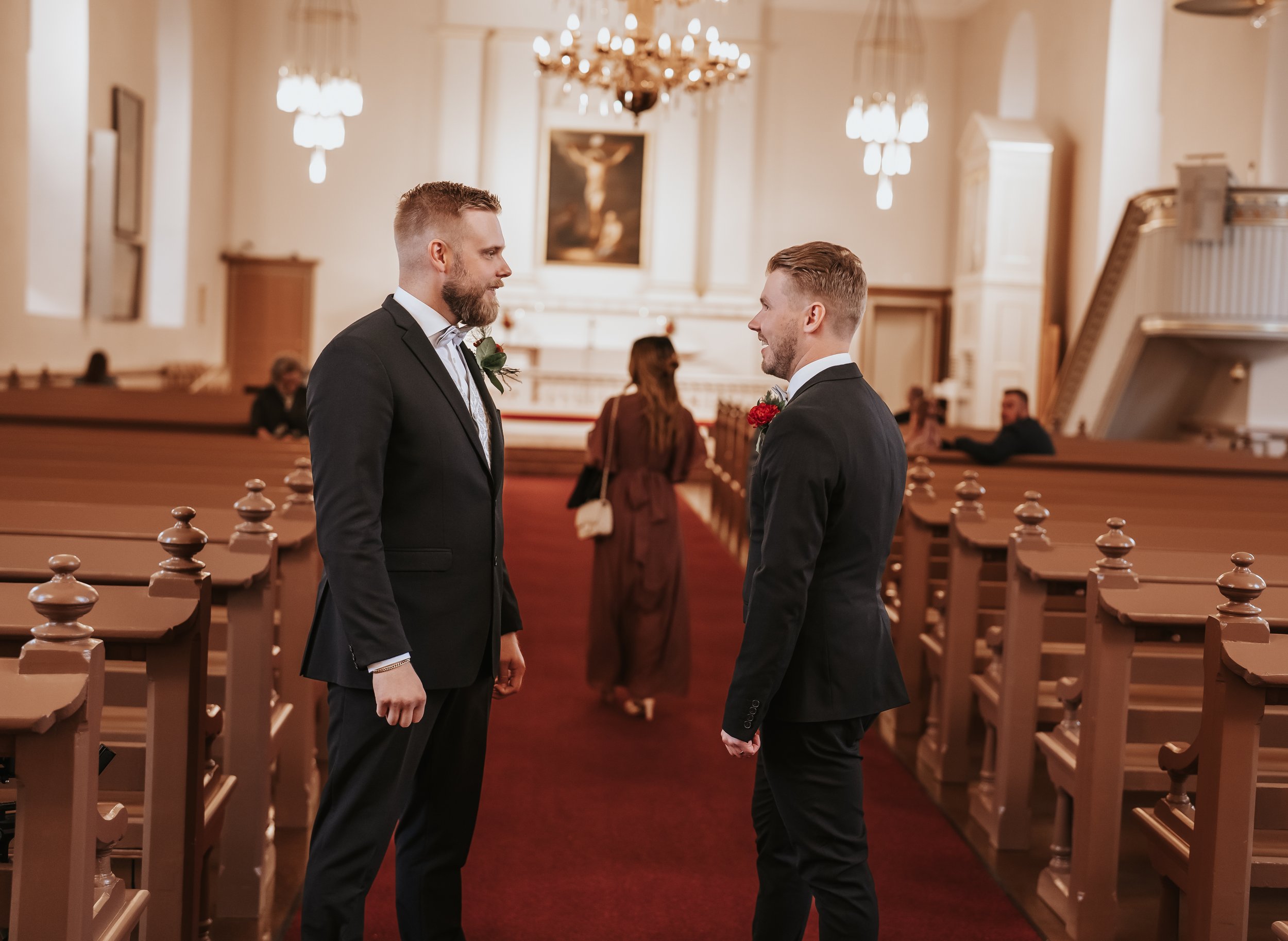 Two men in suits standing in a church aisle, talking before a wedding ceremony, with pews and chandeliers in the background.