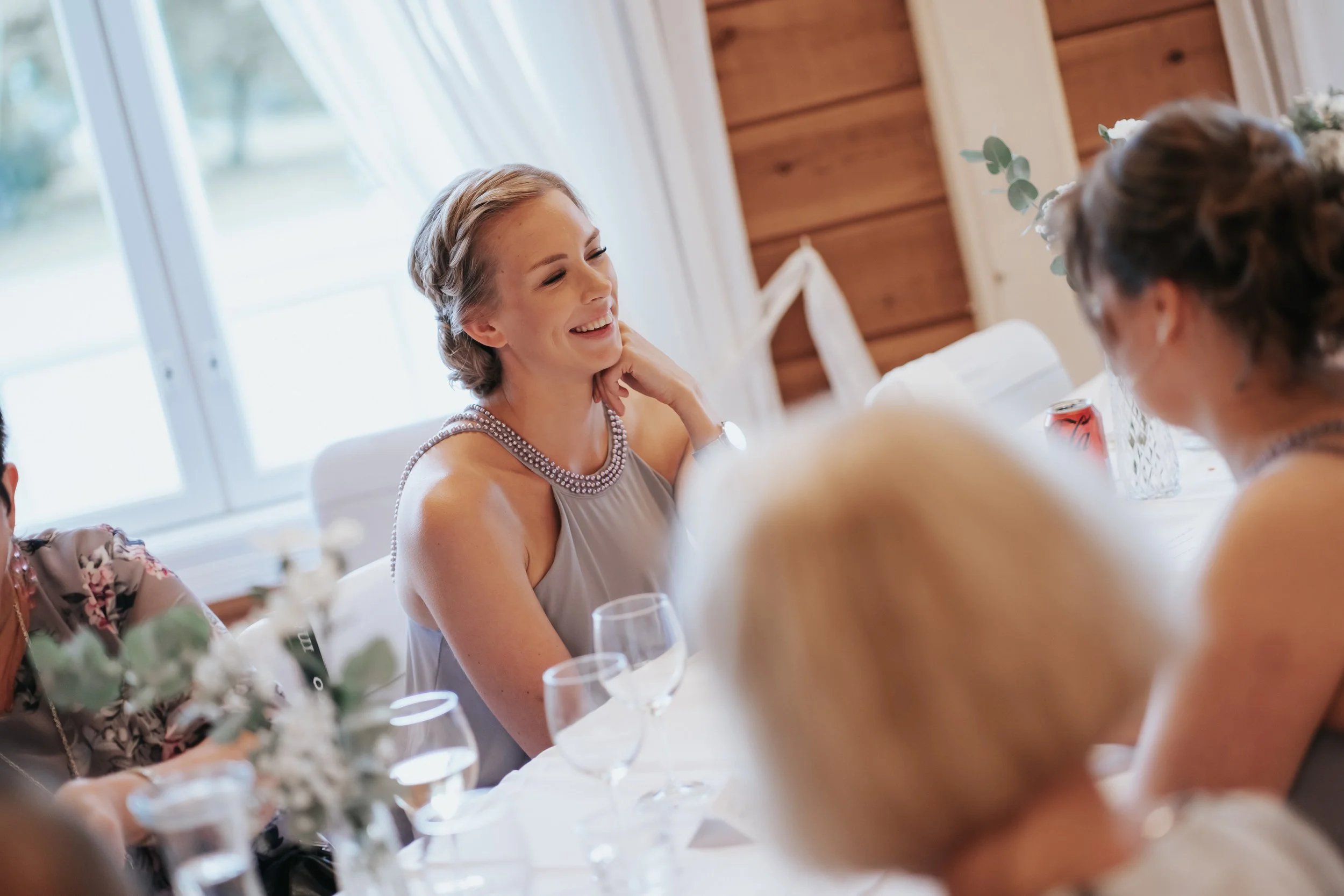 People sitting at a table, a woman smiling, elegant setting, flowers and glassware on the table, window in the background.