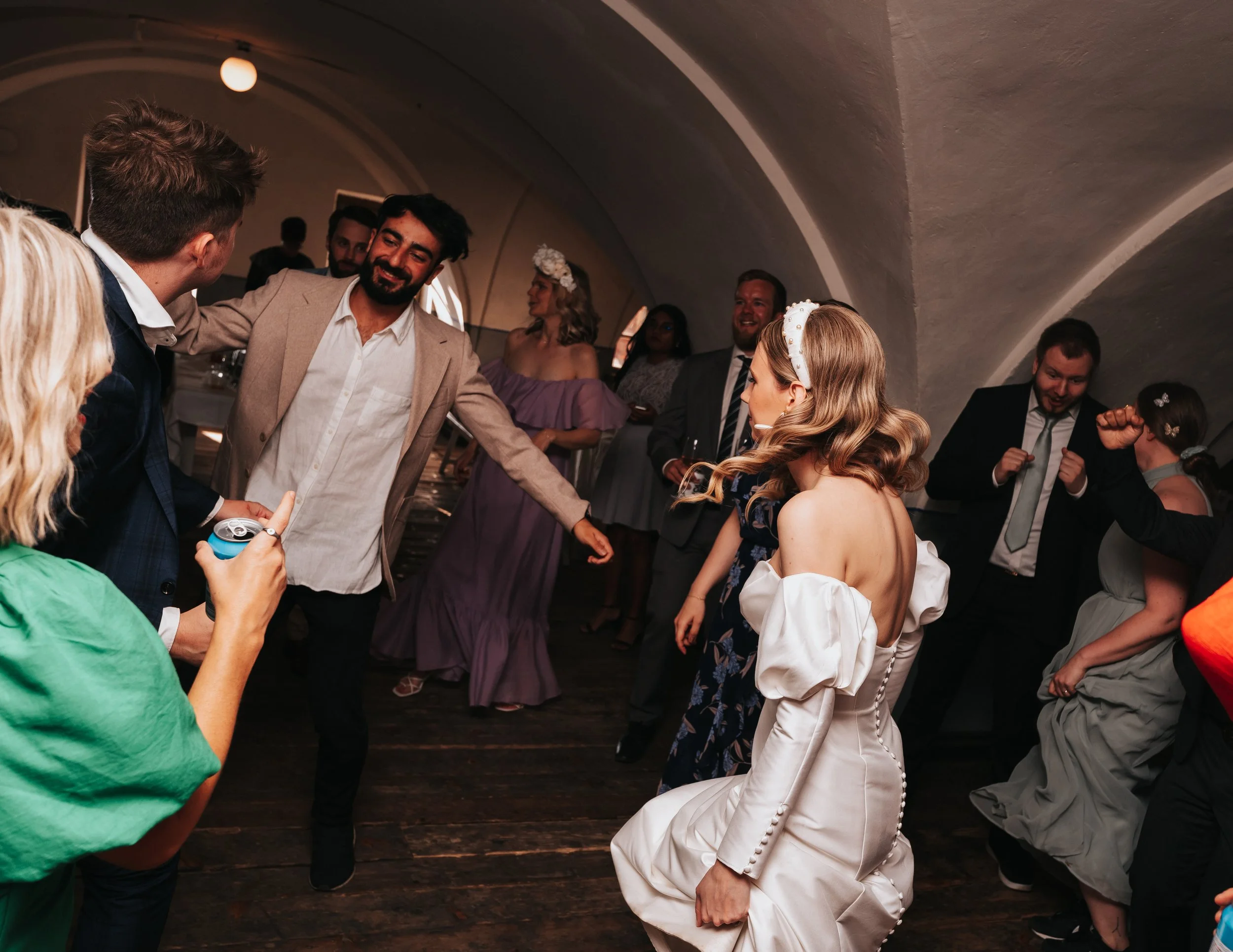 A group of people dancing at an indoor wedding reception. The bride is in a white dress with puffy sleeves, and other guests are in formal attire. The setting is a dimly lit room with an arched ceiling.