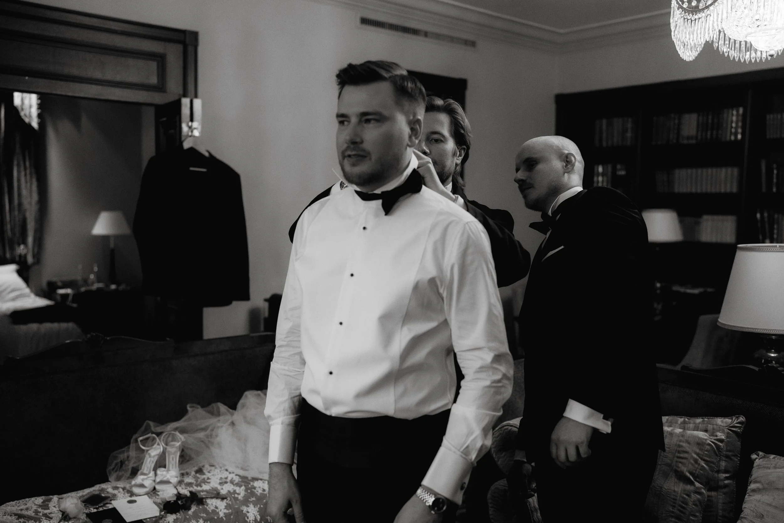 Groomsmen getting ready in a room, dressed in formal attire, with a chandelier and bookshelf in the background.