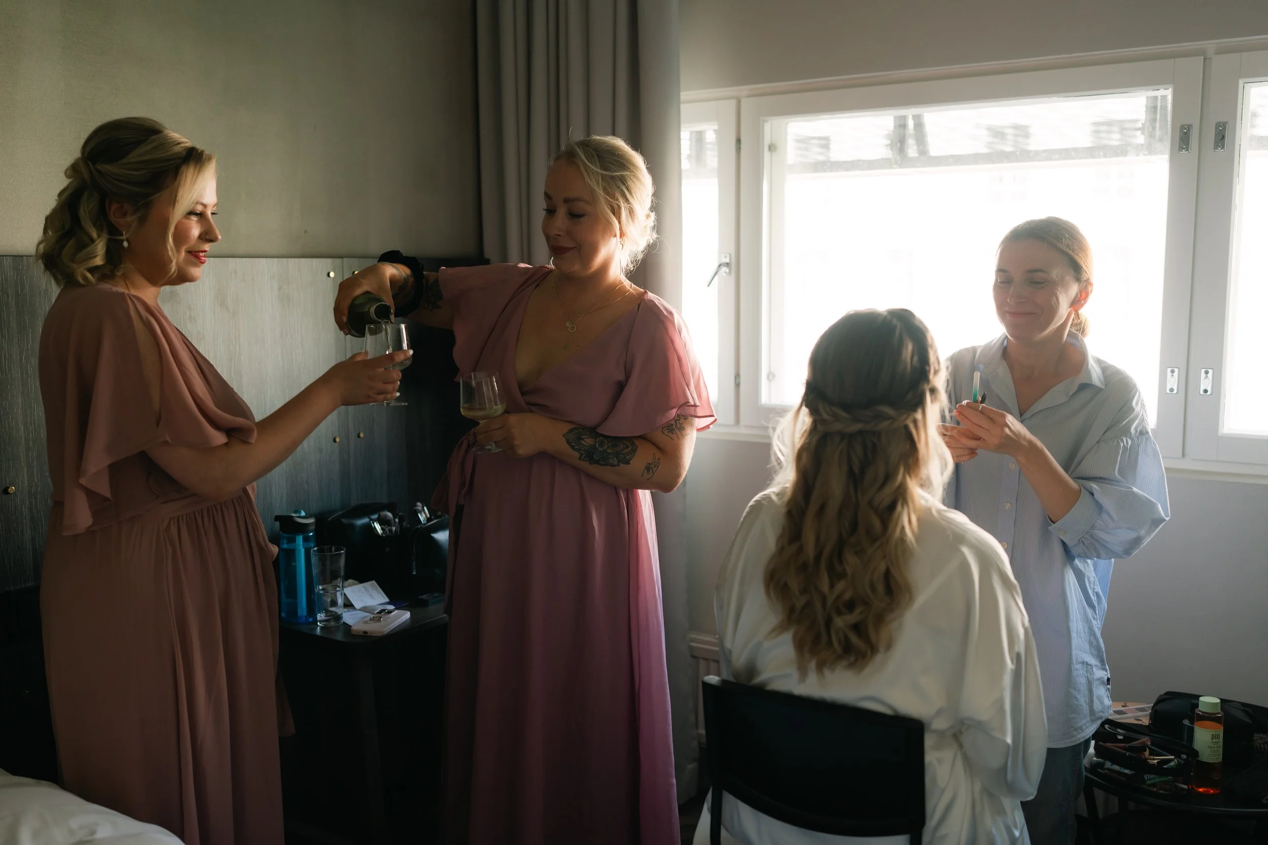 Women in a room, two in pink dresses, holding drinks, and one person getting makeup applied.