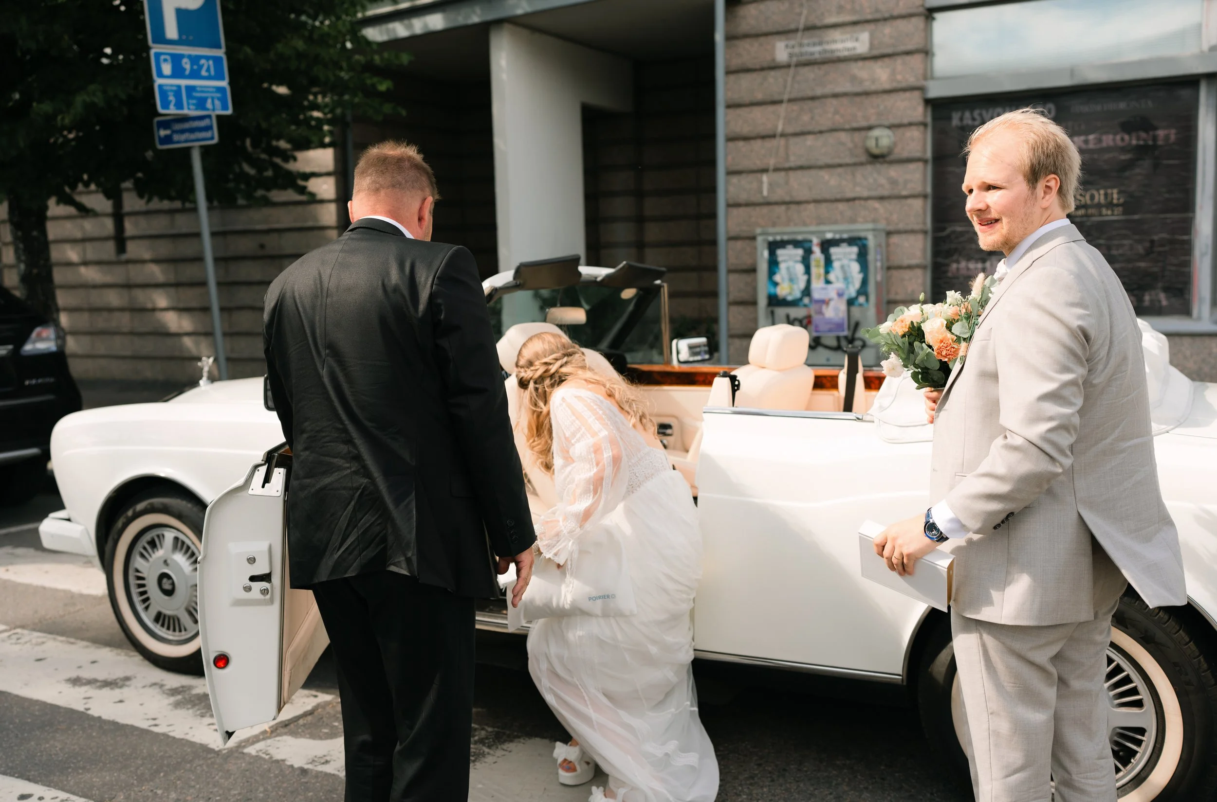 Bride entering white convertible car with two men in suits, holding flowers, outdoor setting.