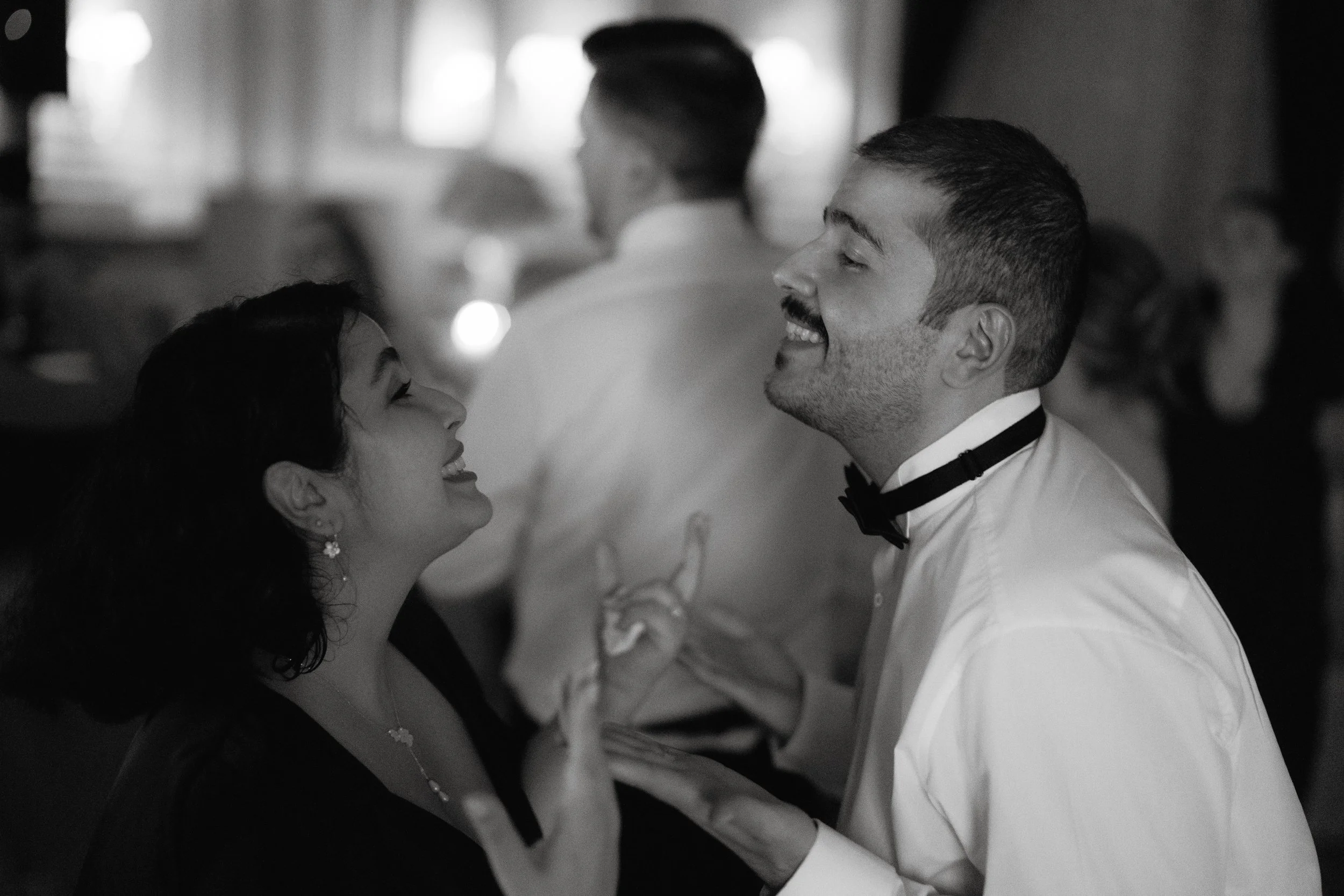 Black and white photo of a man and woman smiling and talking at an event, with another person visible in the background.