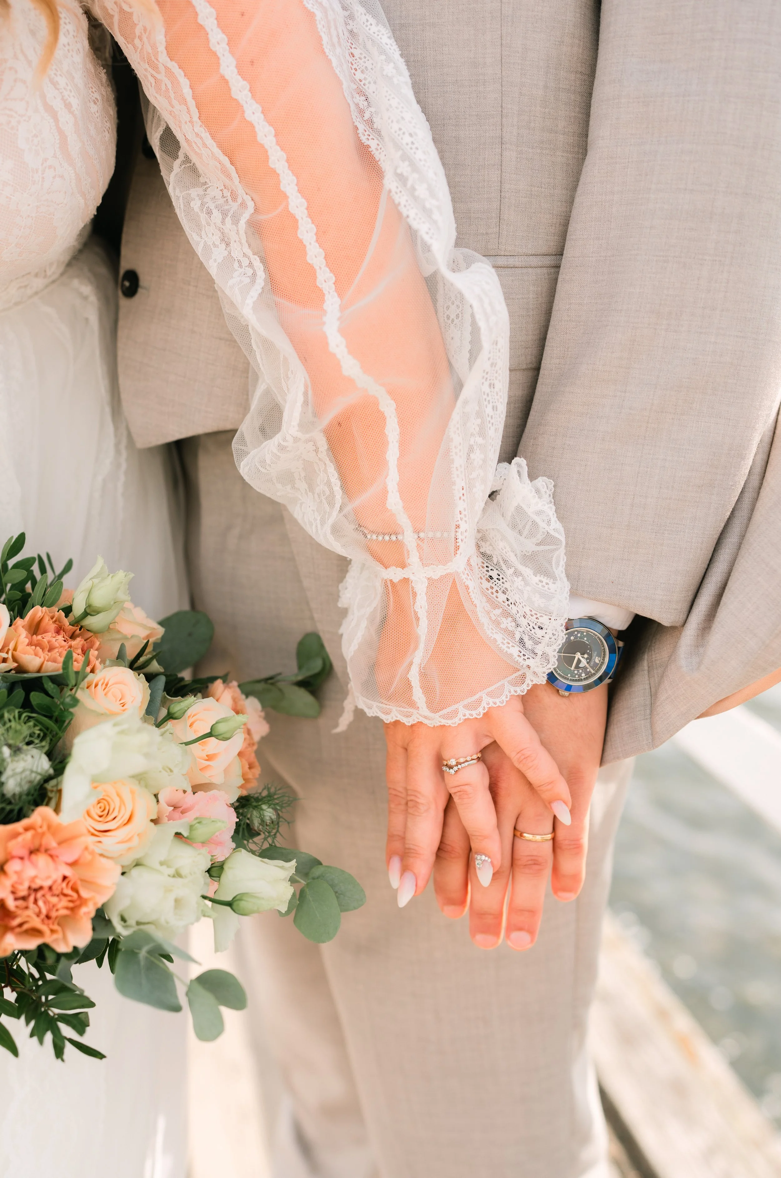 Bride and groom holding hands, bride's lace sleeve, groom's suit, wedding rings, bouquet of roses.