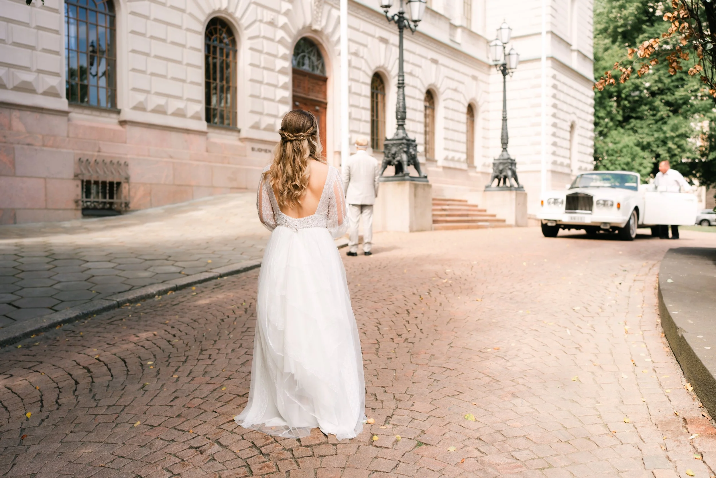 Bride in white gown standing on cobblestone street near vintage car and ornate building.