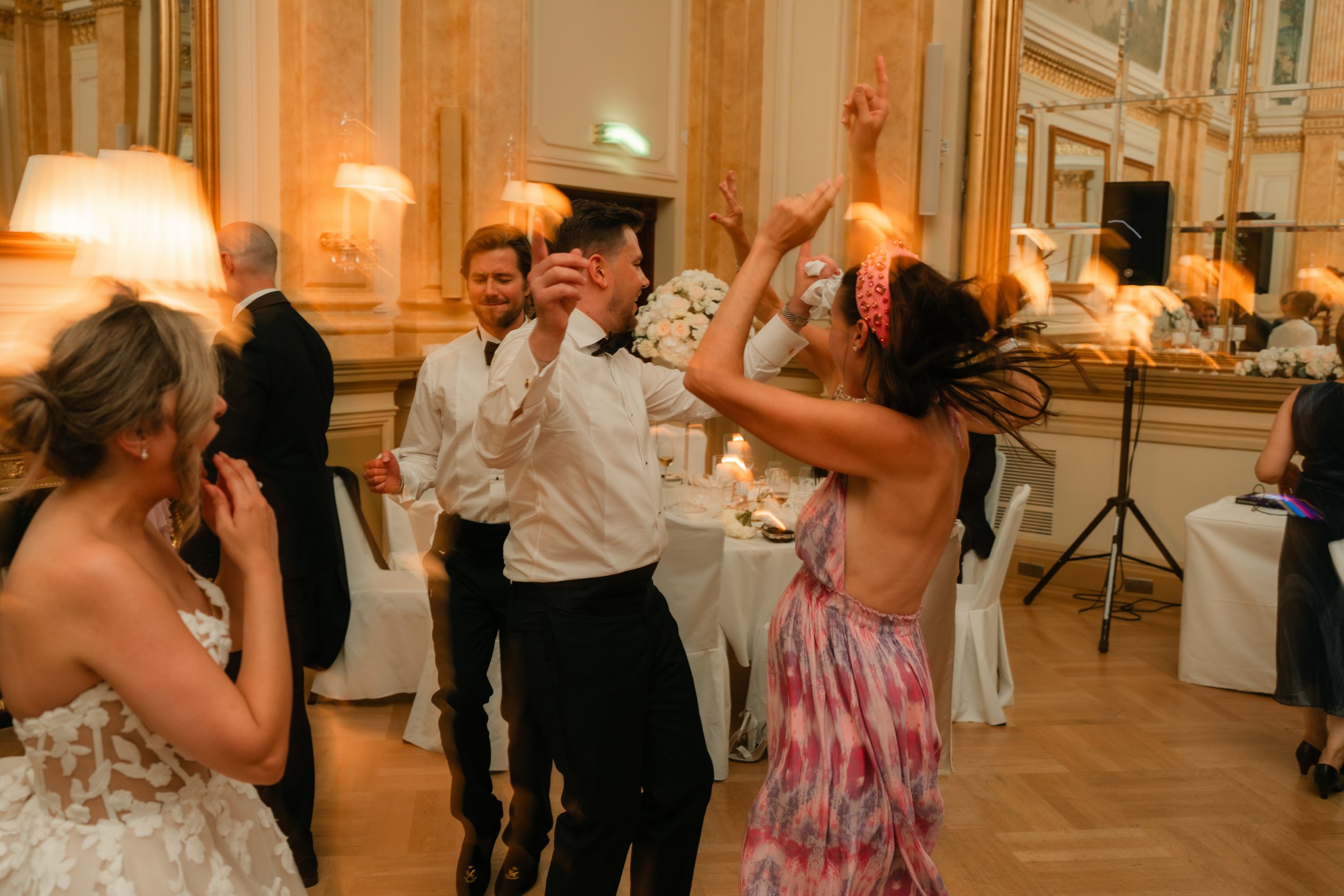 People dancing and celebrating at a formal indoor event, possibly a wedding reception, with elegant decor and blurred motion.