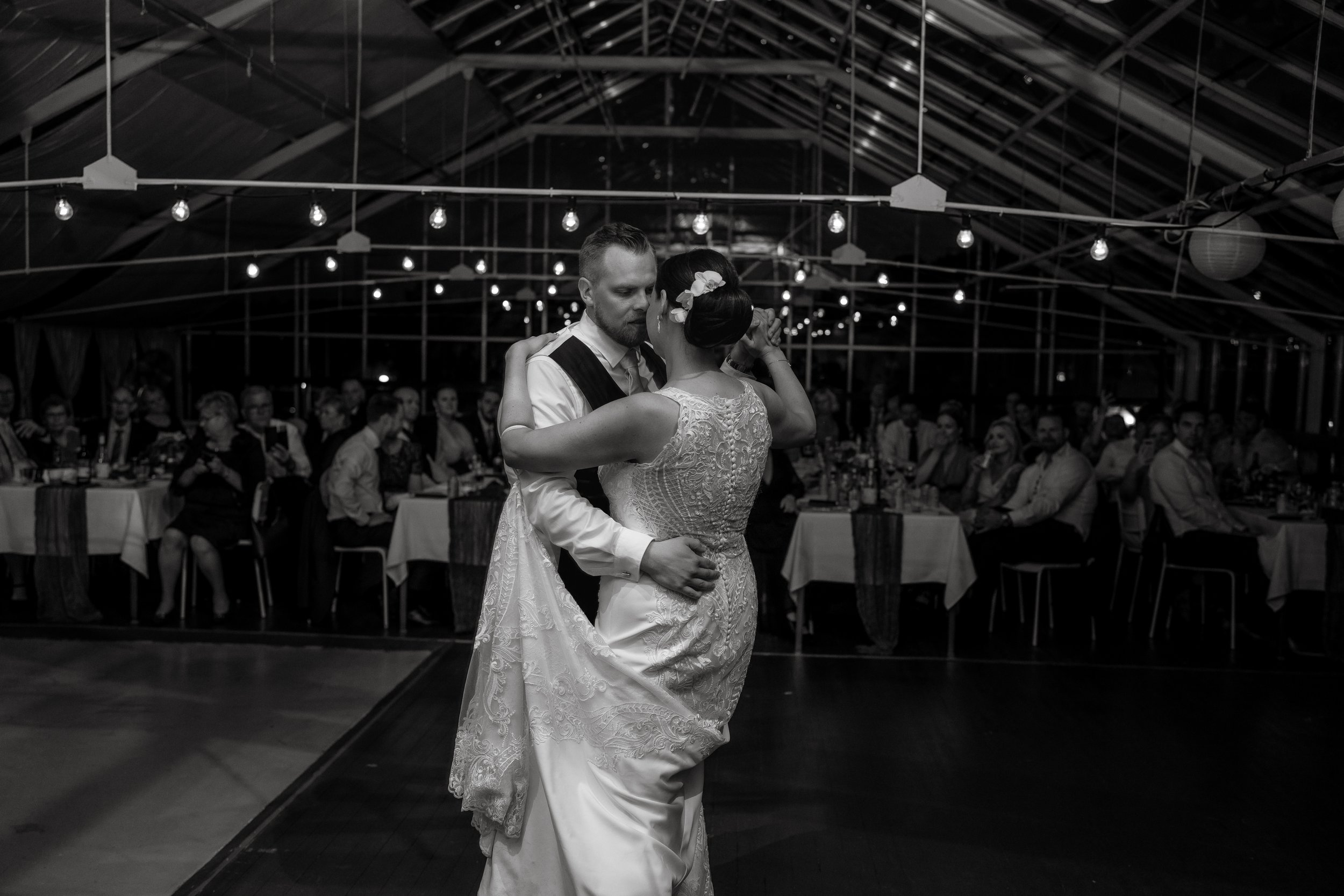 Bride and groom dancing at wedding reception, black and white photo, guests seated and watching, string lights overhead.