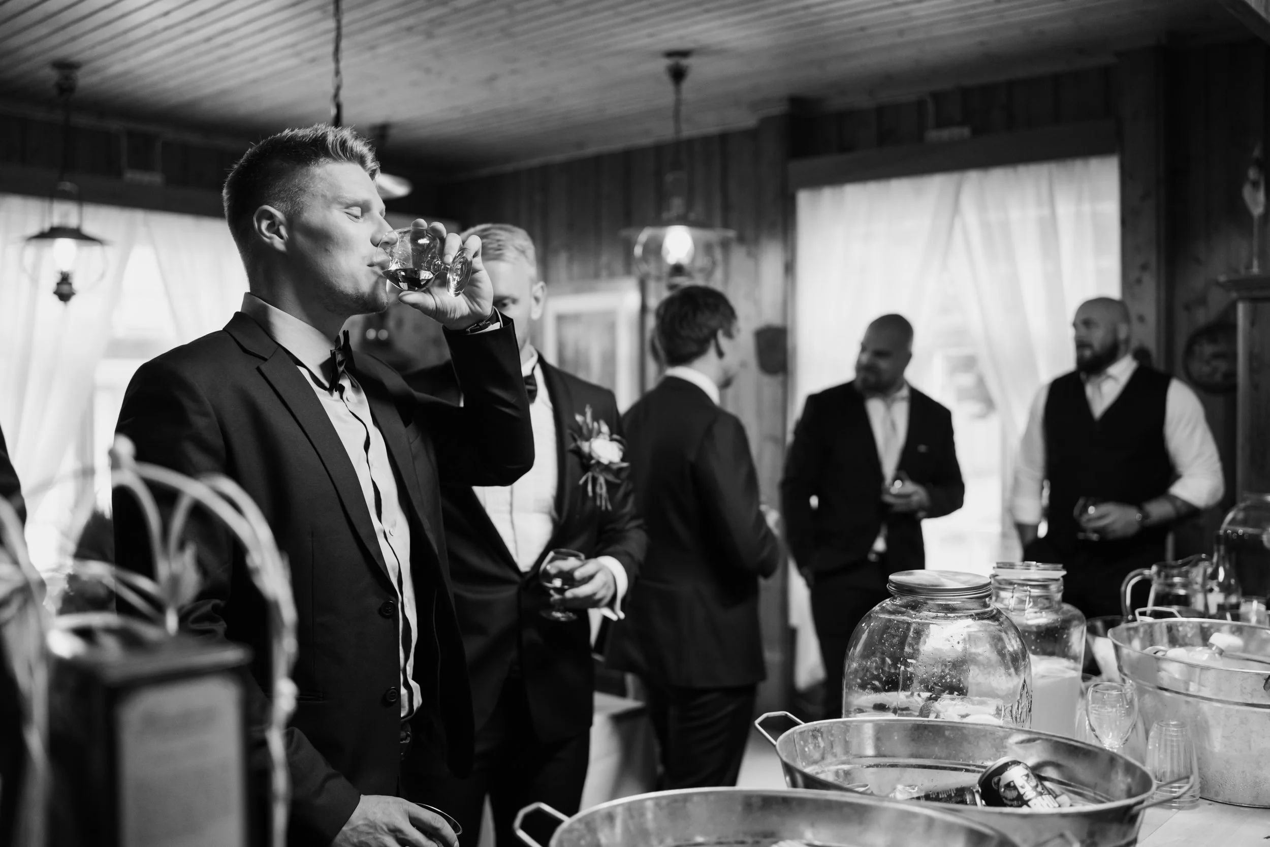 Black and white image of men in suits drinking at a social gathering, with ice buckets and jars in the foreground.