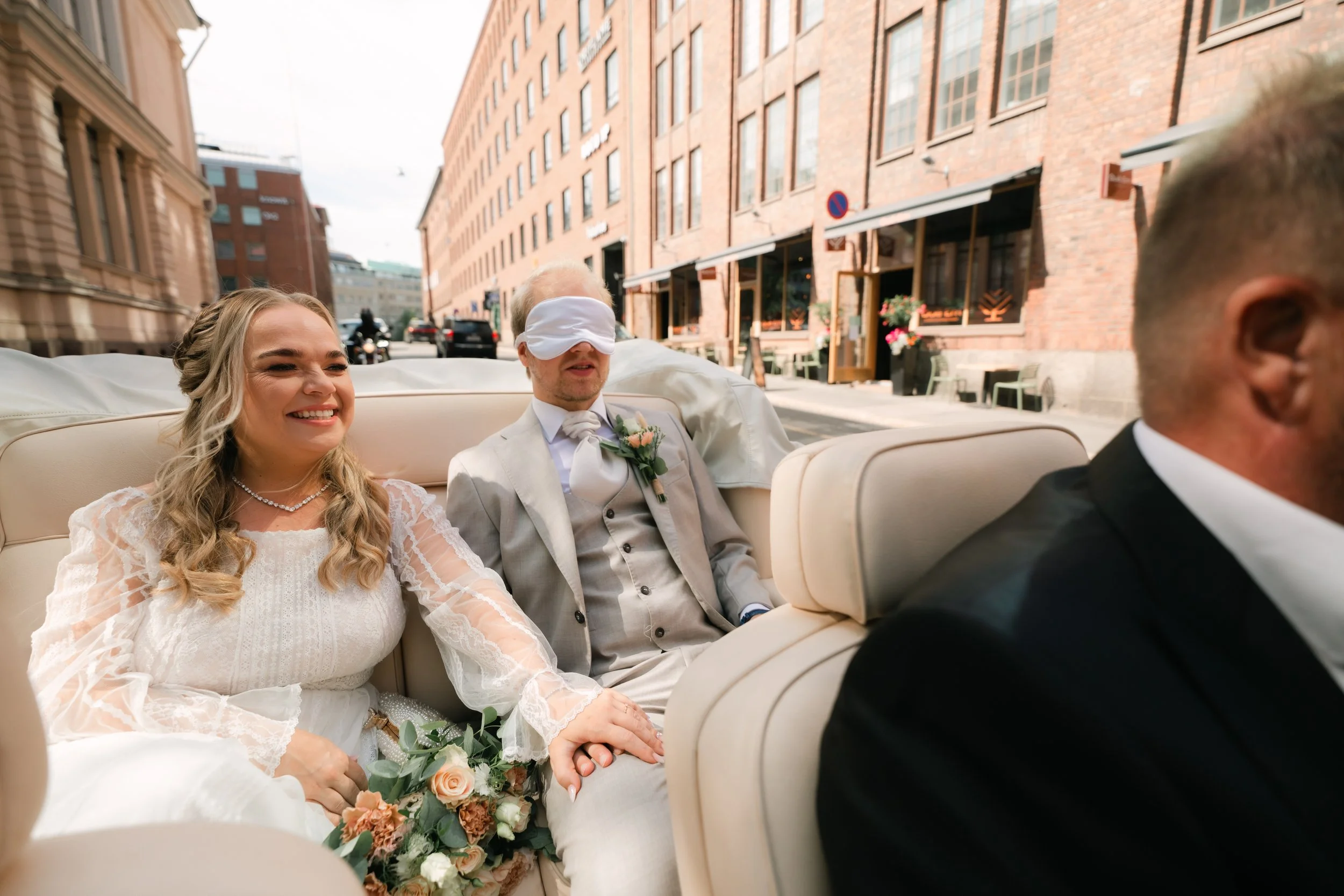 Bride and groom in a convertible car, groom blindfolded, smiling bride holding bouquet, urban background.