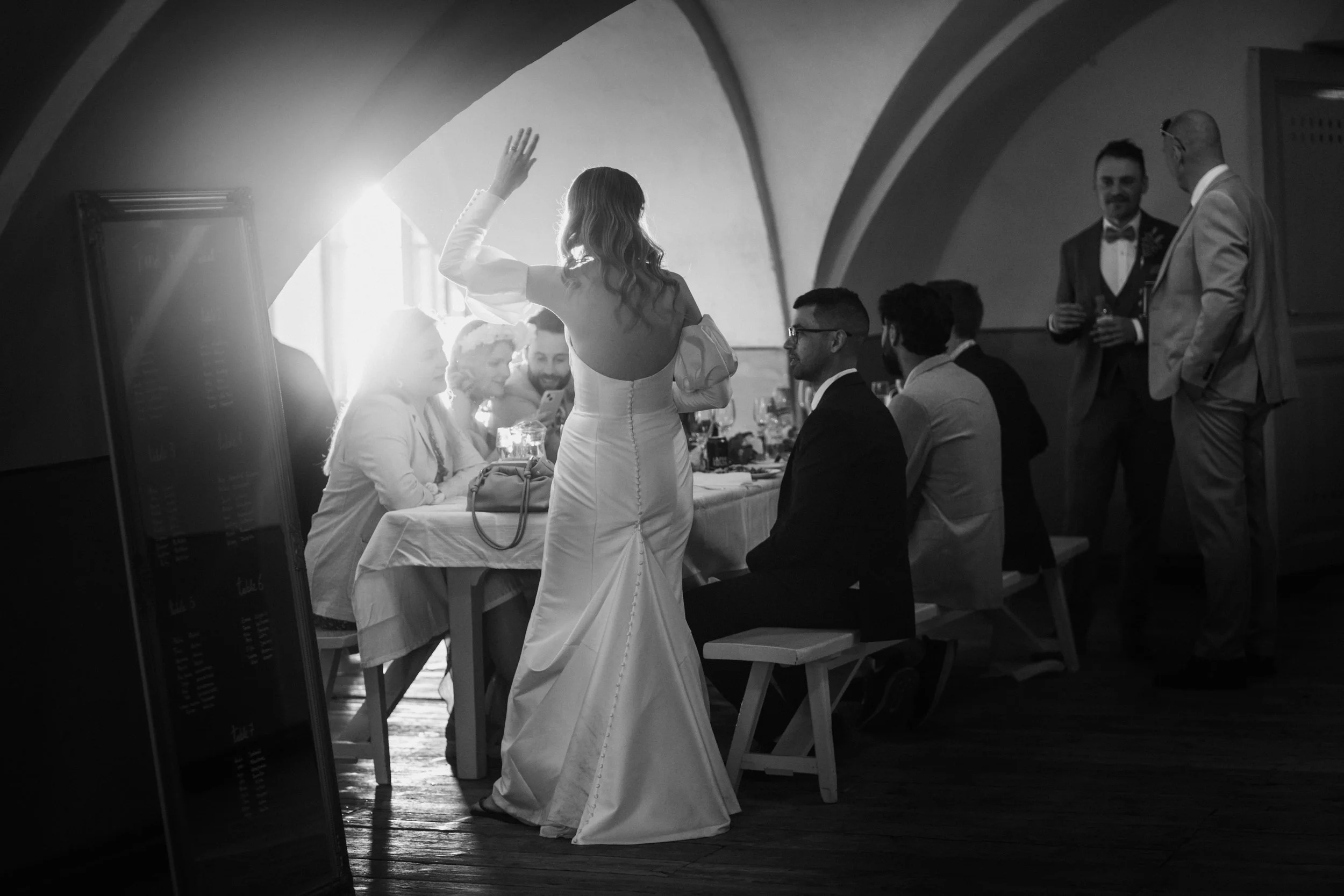 A black and white photo of a wedding scene with people seated at a table. The bride, in a wedding dress, stands with her back to the camera, raising her hand. Guests are sitting and standing around her, conversing. Sunlight streams in from a window.
