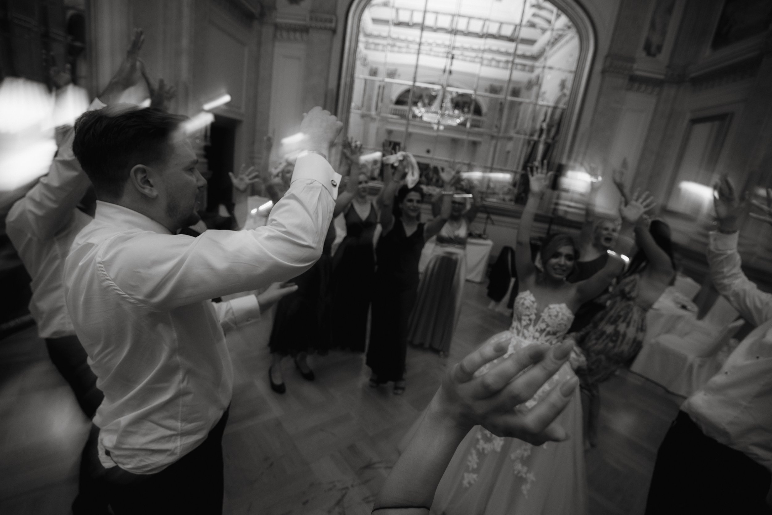 Black and white photo of people dancing indoors with hands raised, wearing formal attire.