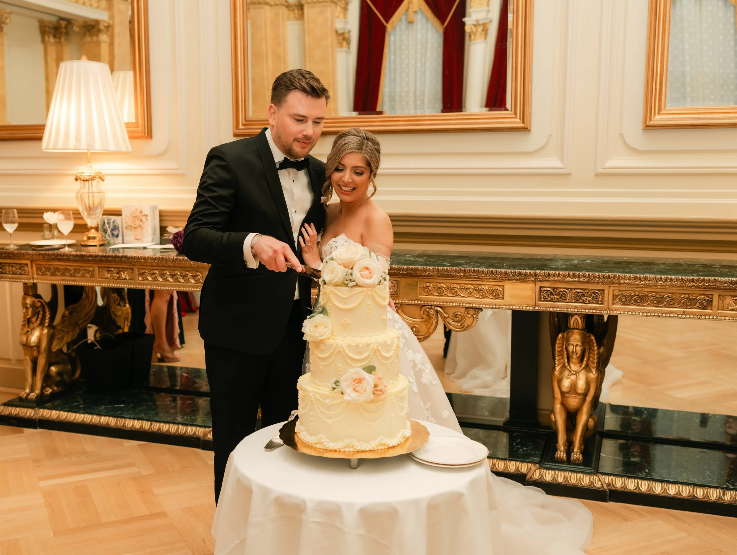 Bride and groom cutting wedding cake in elegant room with ornate decor and mirrors.