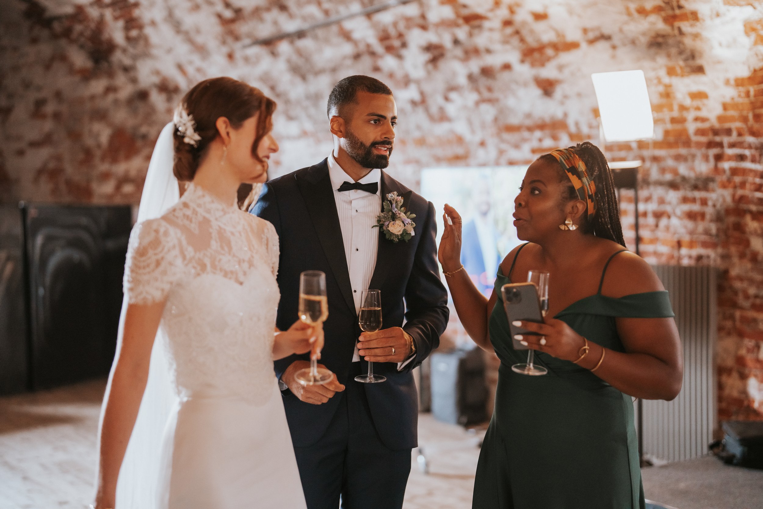 Bride, groom, and guest at wedding reception, indoor venue, holding champagne glasses.