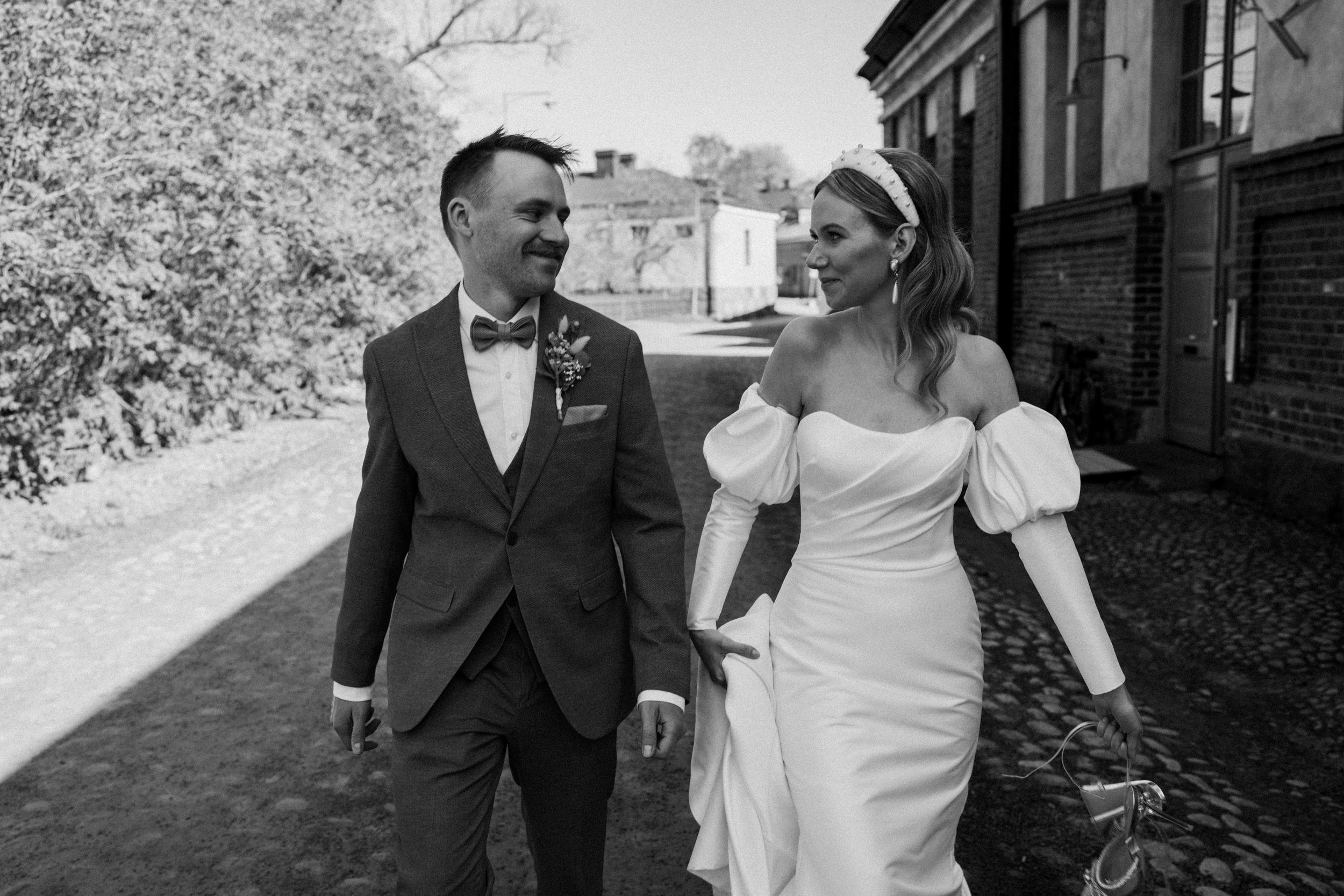 Bride and groom walking outdoors, smiling at each other, in black and white photo.