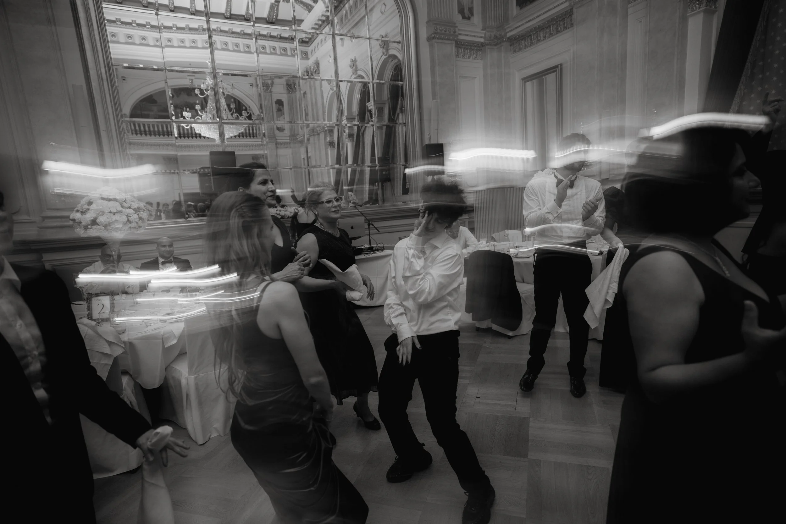Black and white photo of people dancing at an indoor event with a mirrored wall and ornate ceiling.