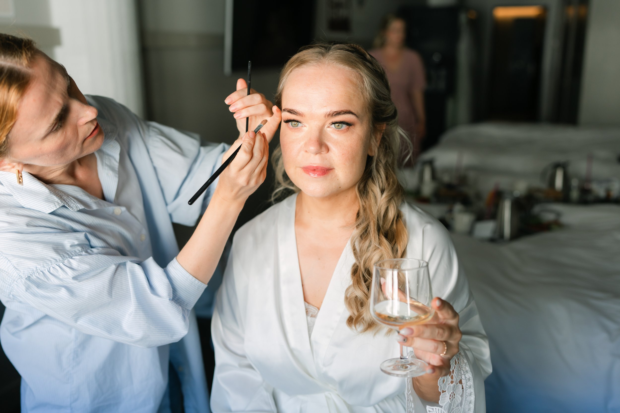 Woman having makeup applied by artist while holding wine glass.