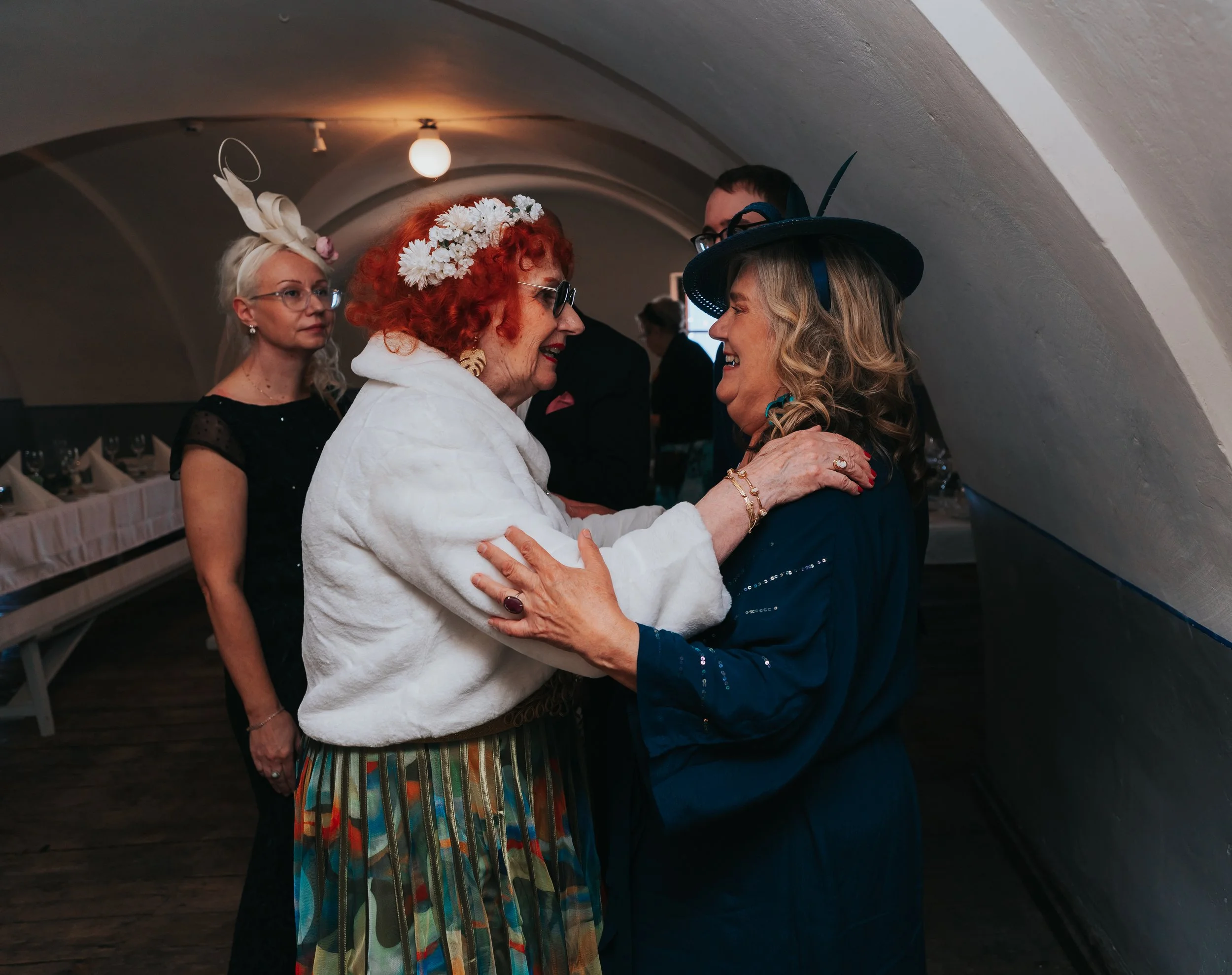 Group of people at a social event, some in flamboyant hats, greeting each other warmly.