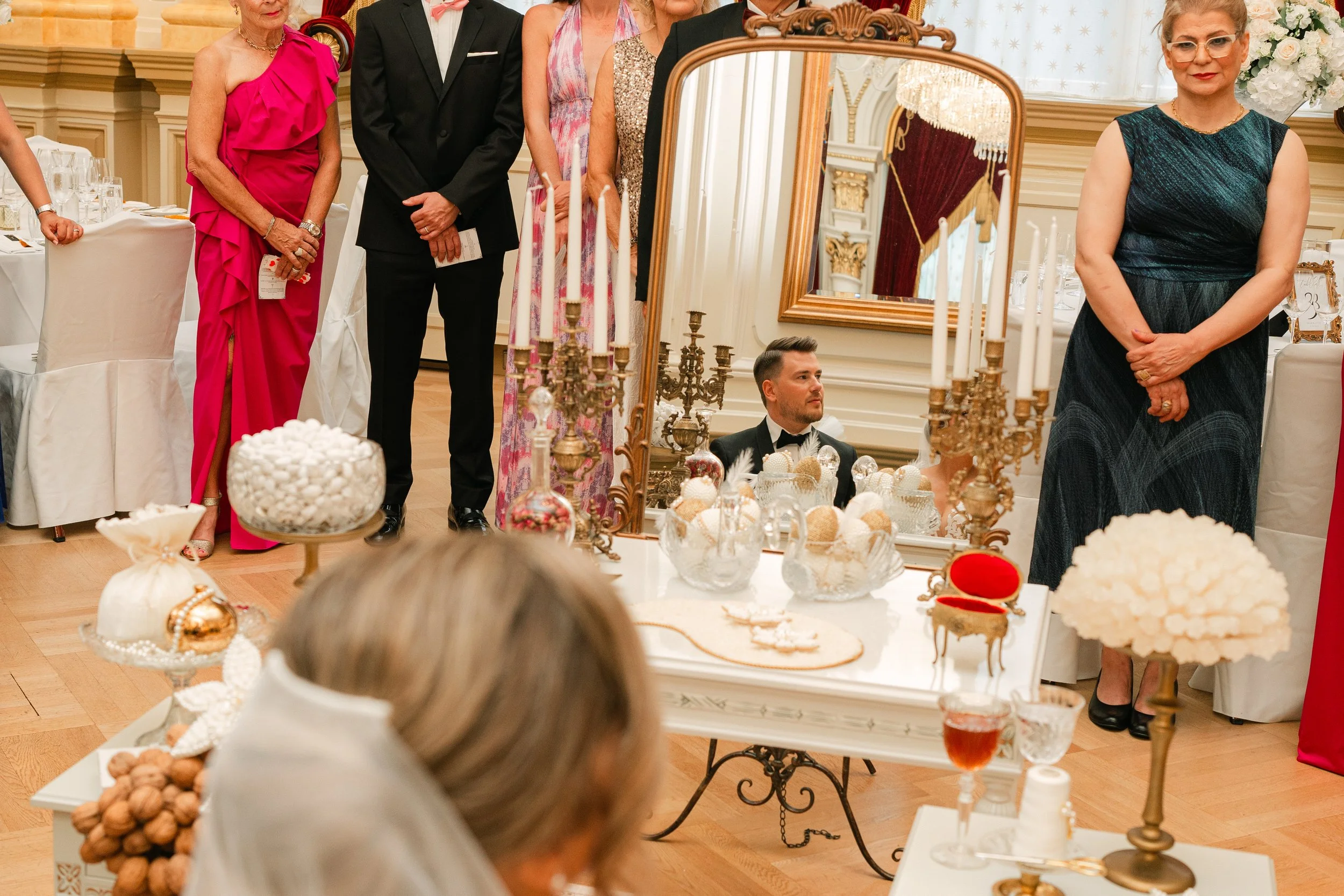 Elegant celebration scene with a table display including candles, decorative bowls, and confections, surrounded by formally dressed guests, reflecting sophistication and opulence in a luxurious setting.