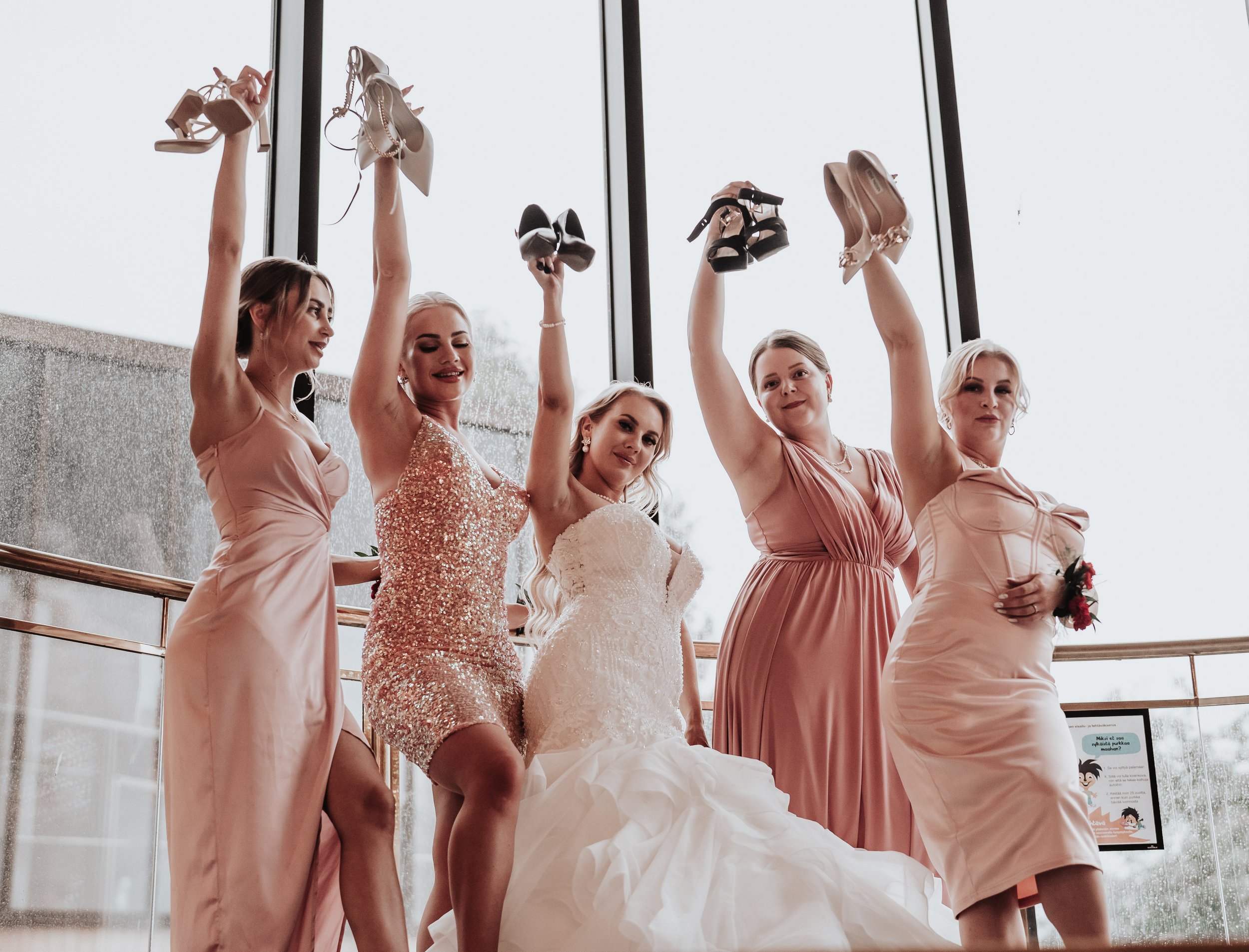 Bridal party posing with bride and bridesmaids holding shoes indoors.