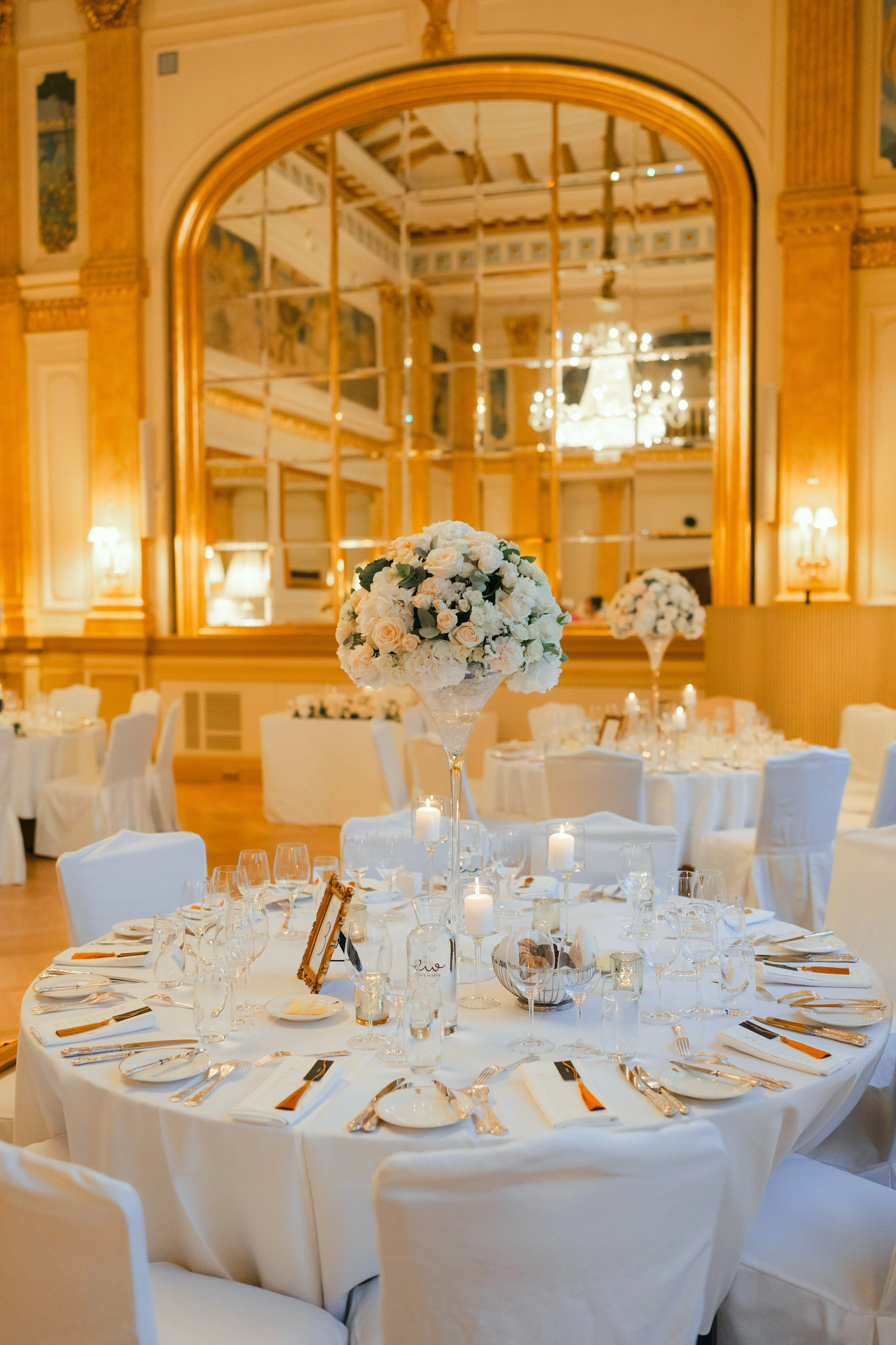 Elegant wedding reception hall with decorated tables, white tablecloths, floral centerpieces, and warm lighting.