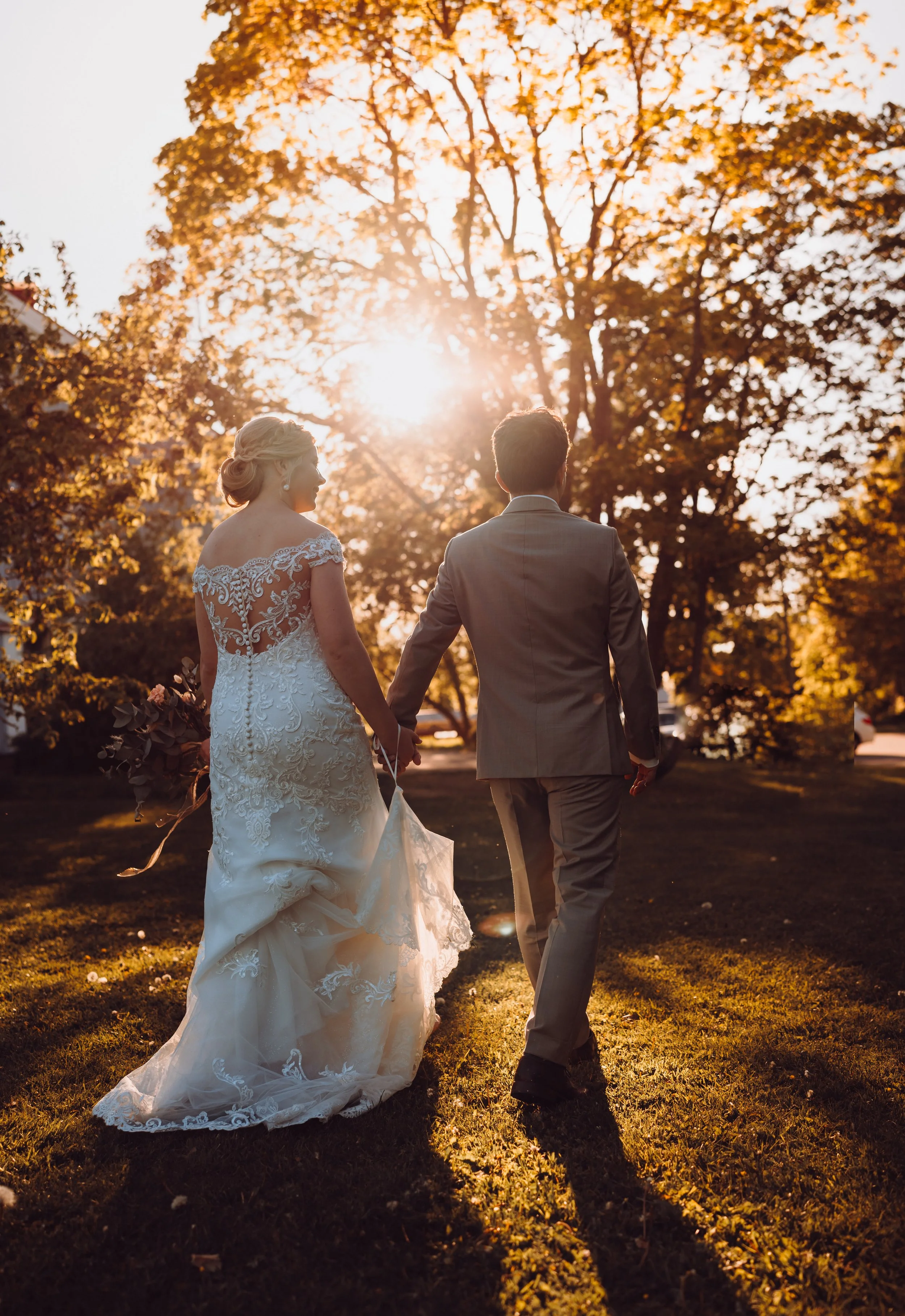 Bride and groom walking hand in hand, backlit by setting sun, in a park with autumn trees.
