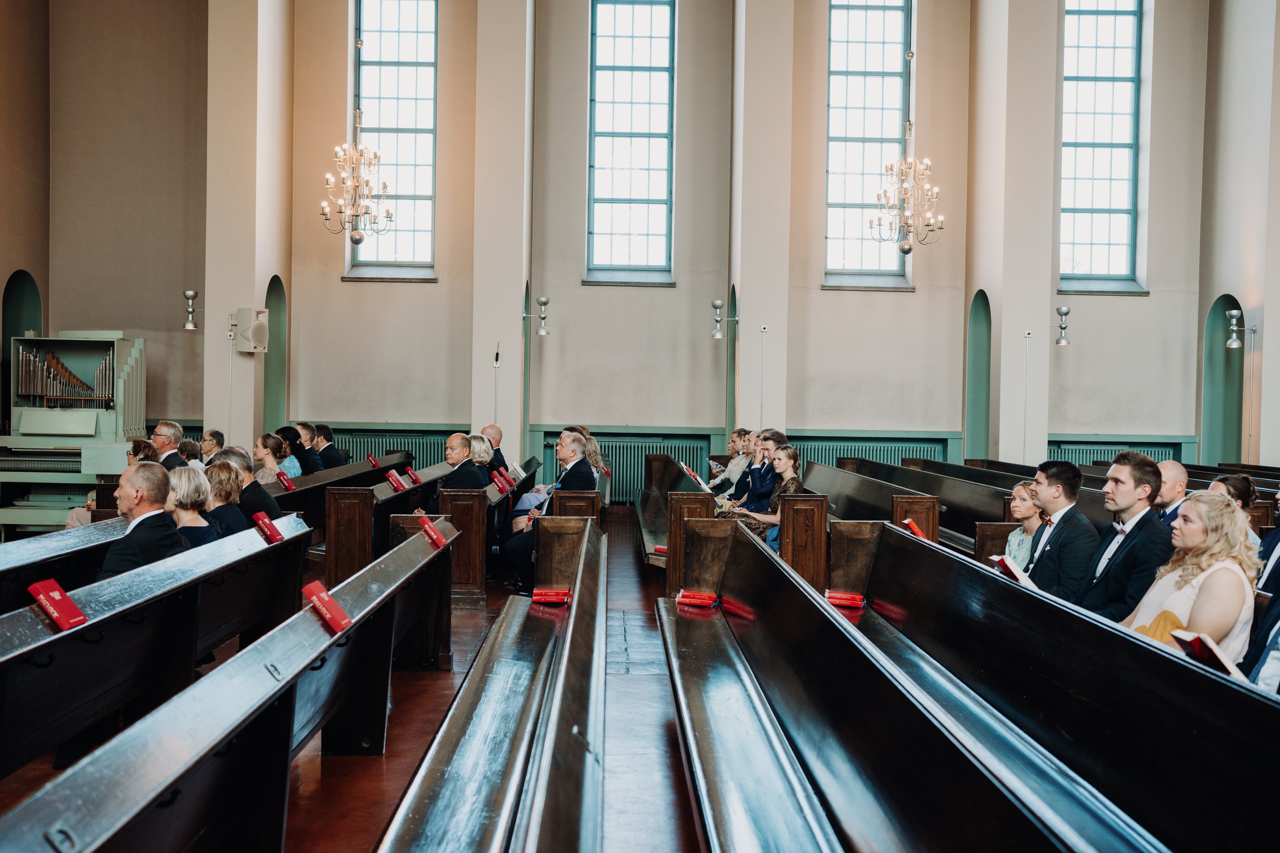 People sitting in church pews during a service, with chandeliers and windows in the background.