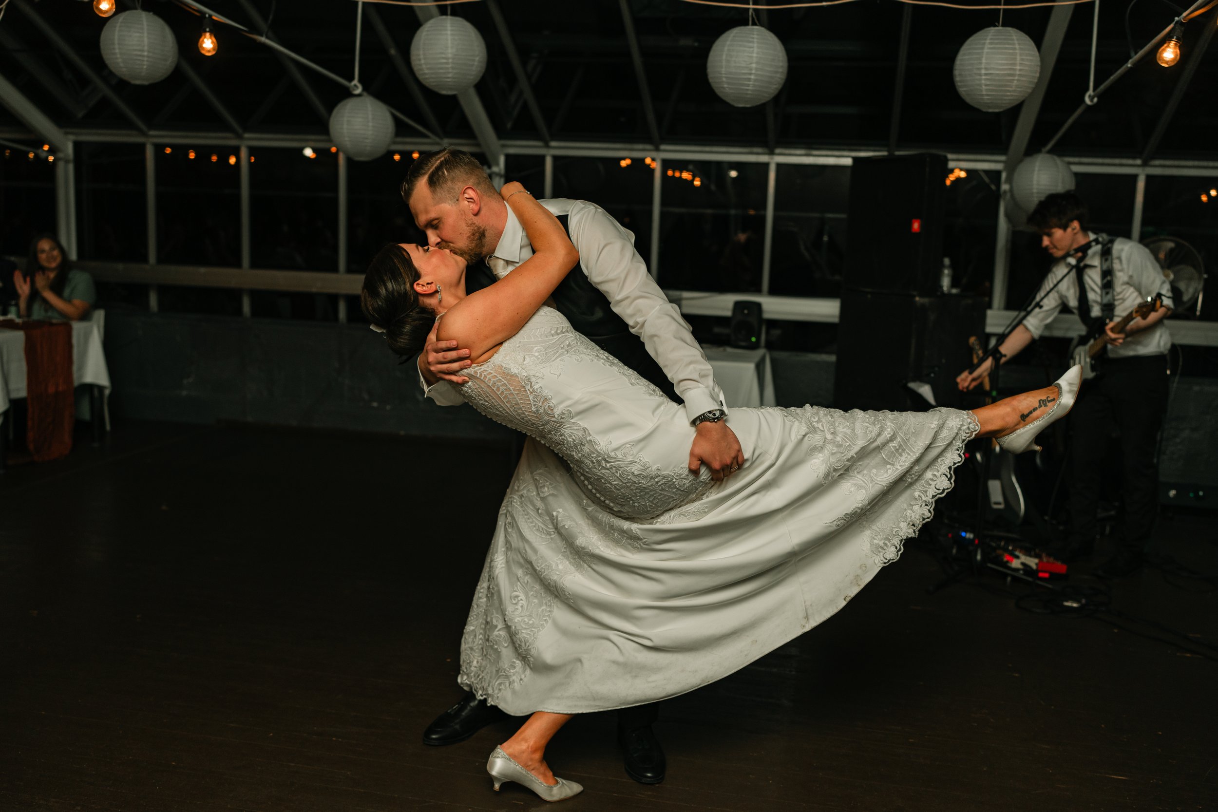 Couple dancing at wedding, groom dipping bride, indoor venue, hanging lanterns, guitarist in background.