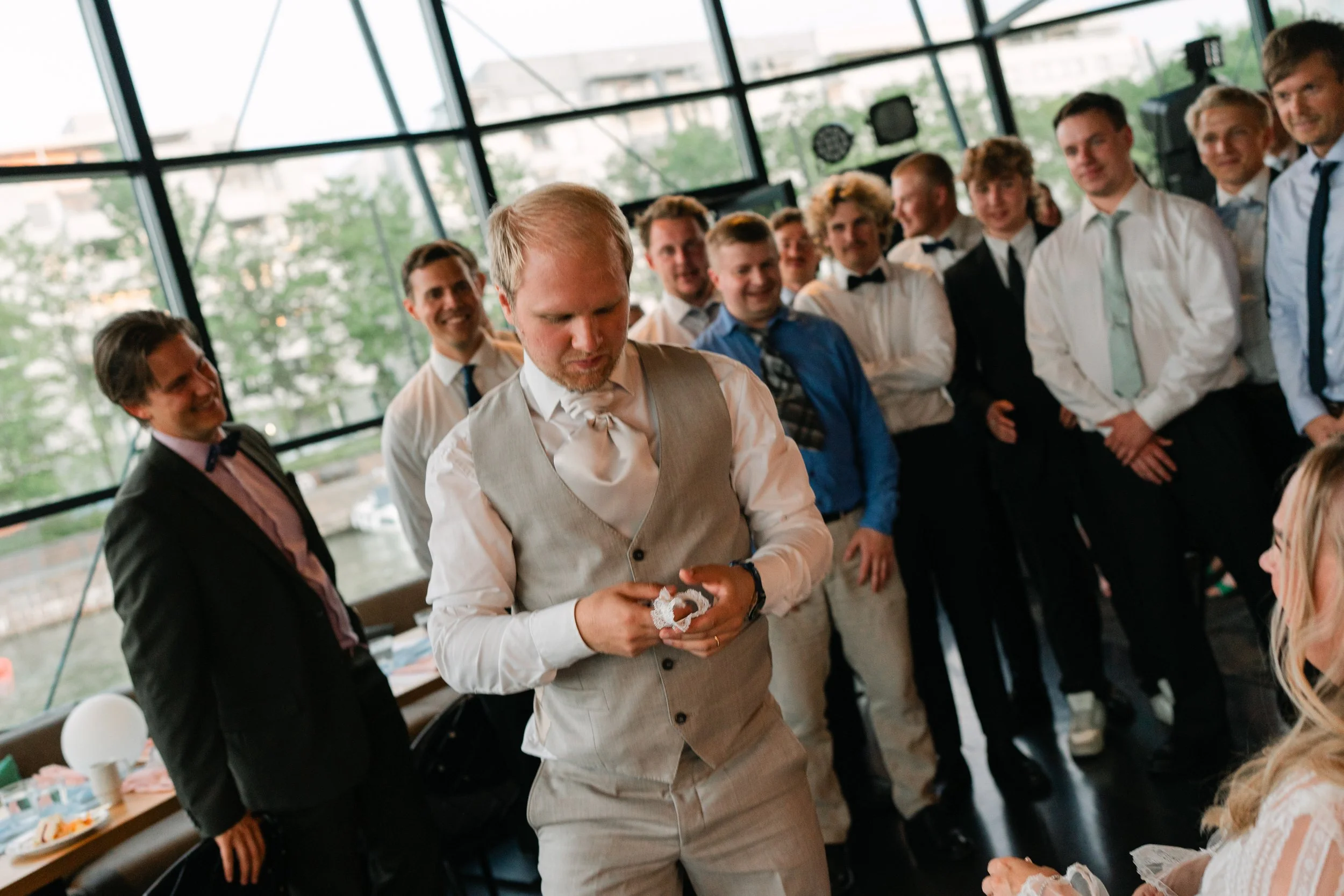 Wedding scene with groom holding garter, surrounded by smiling men, indoor setting with large windows.