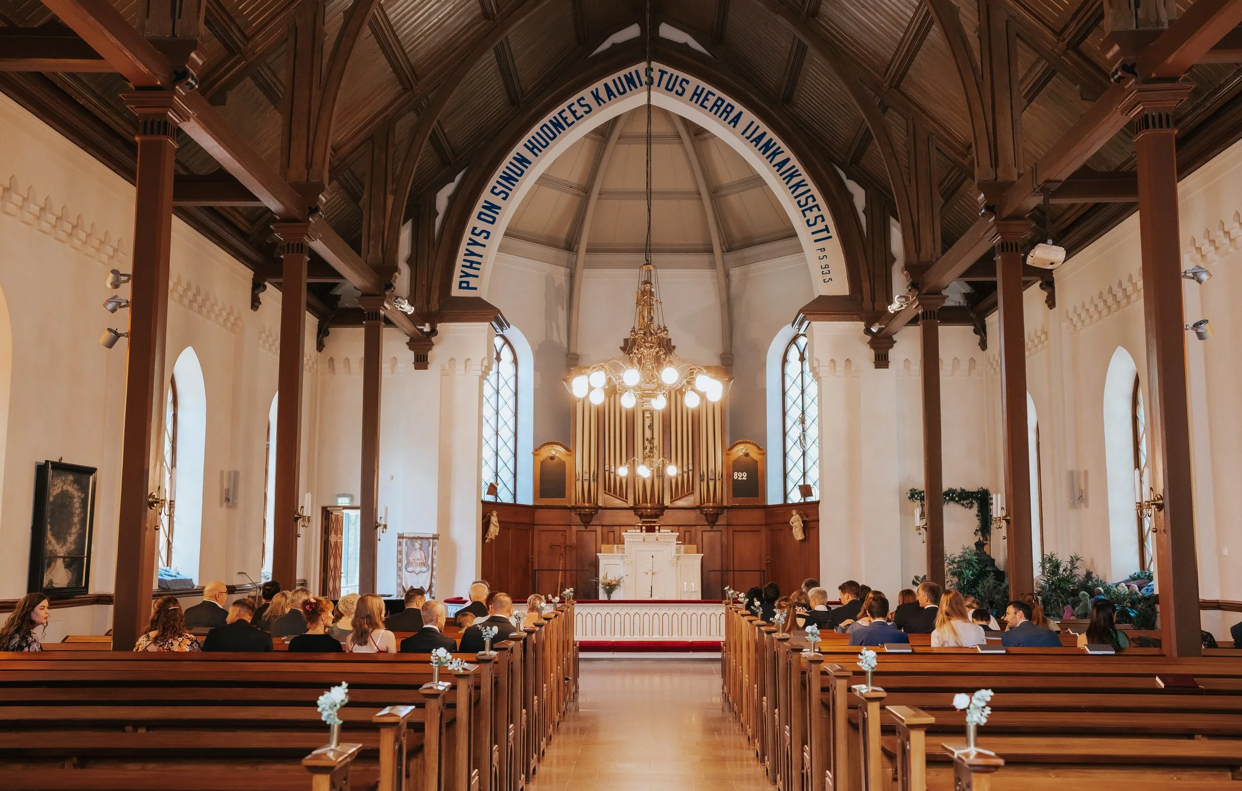 Interior of a church with wooden pews and altar, people seated, large organ and chandelier, stained glass windows, Finnish text on arch.