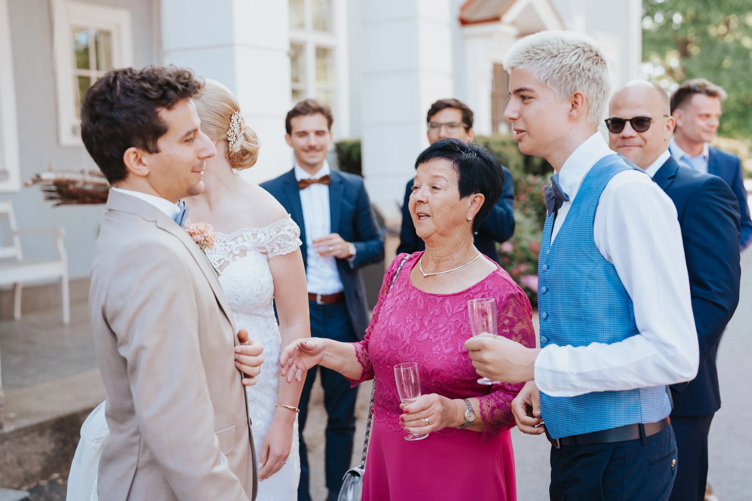 Group of people in formal attire at an outdoor event, including a bride in a wedding dress and guests holding champagne glasses.