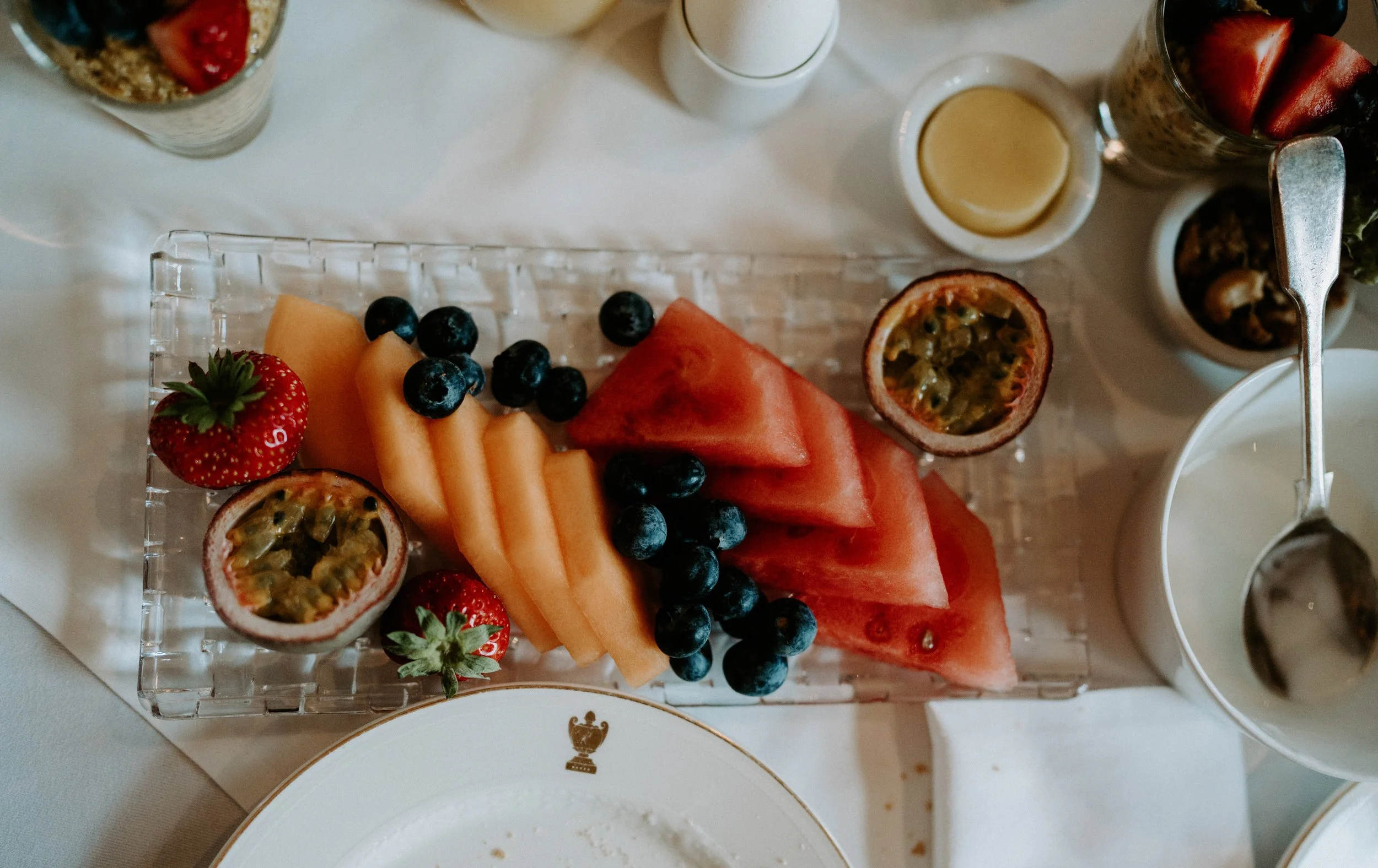 Platter of sliced watermelon, cantaloupe, blueberries, strawberries, and passion fruit on a table with yogurt and other breakfast items.