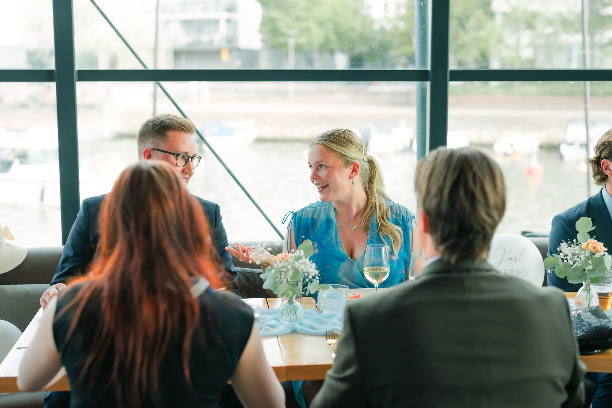 People sitting at a table indoors, talking and smiling, with flowers and drinks on the table, near large windows with a waterfront view outside.