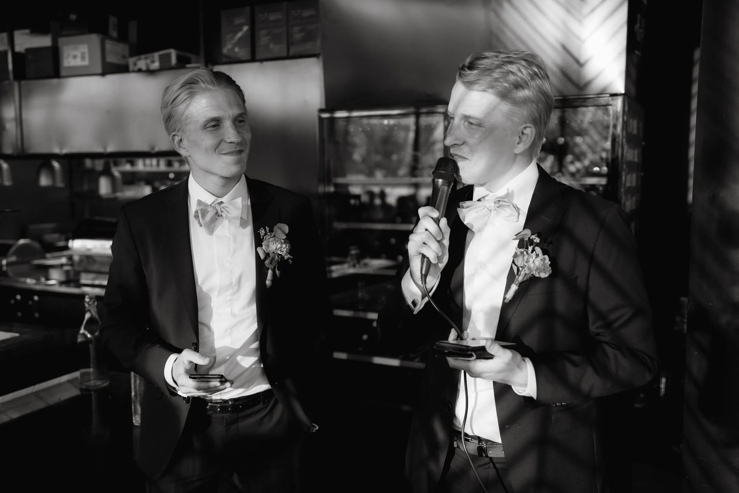Two men in suits with boutonnieres speaking at an event, one holding a microphone.