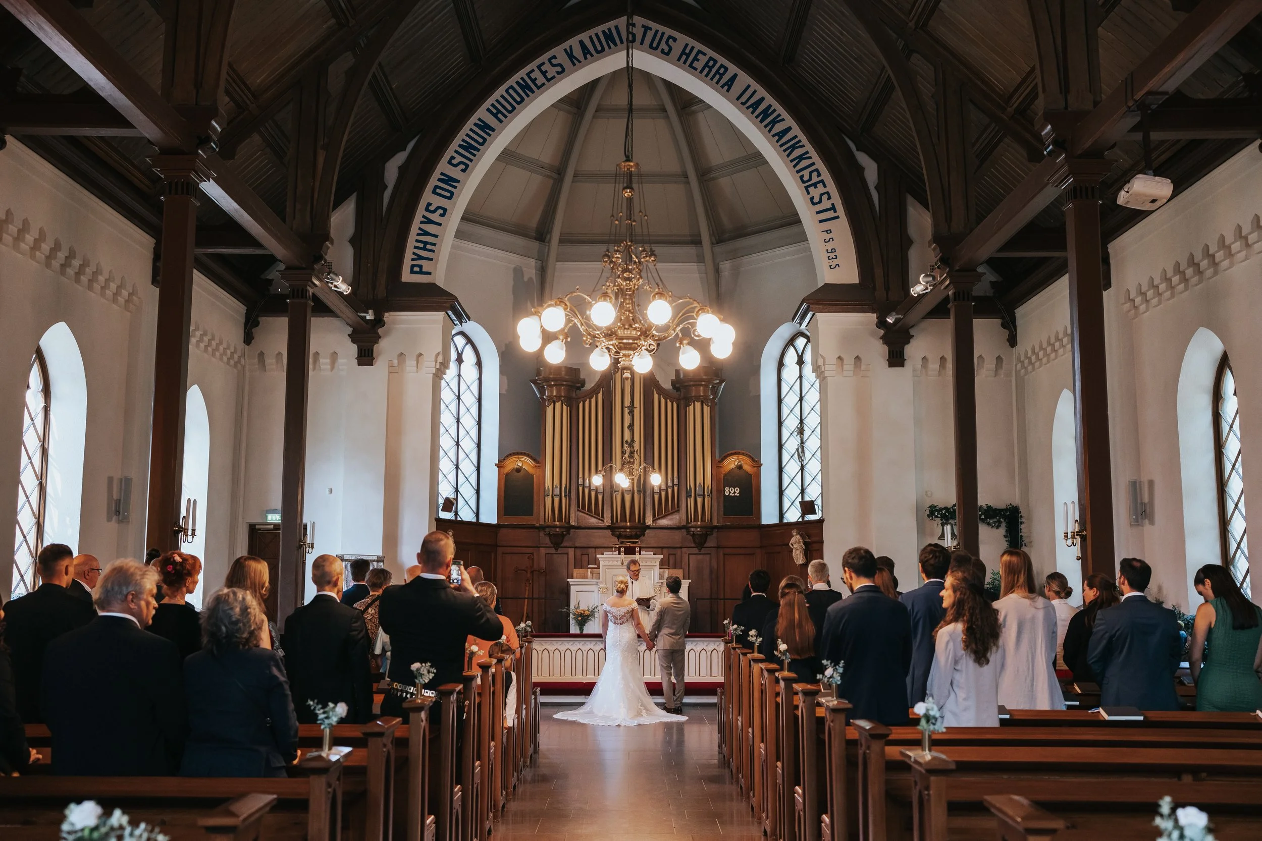Interior of a church during a wedding ceremony with a couple standing at the altar, guests seated on wooden pews, and an organ in the background; Finnish text on the archway.