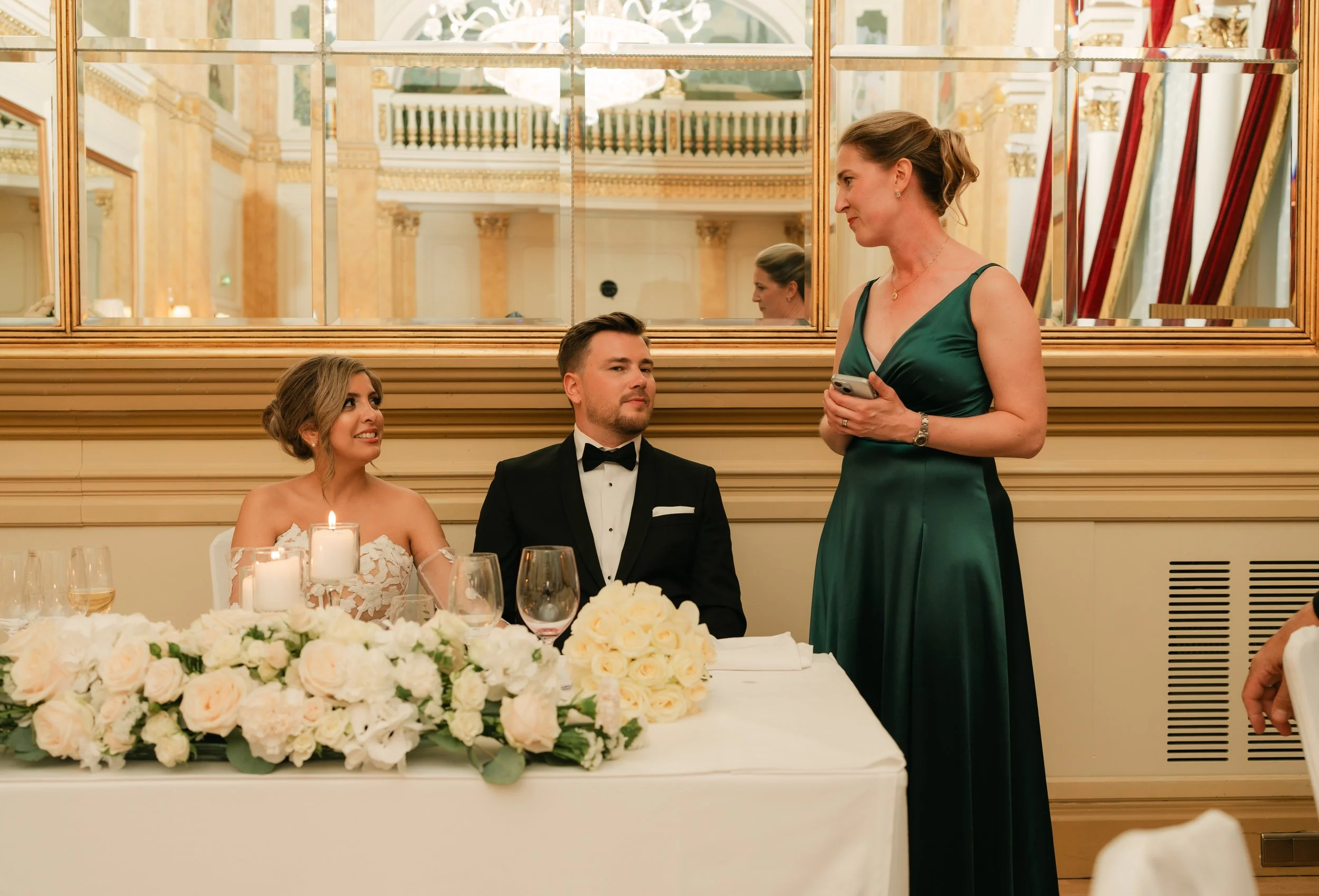 Wedding scene with bride and groom seated at a table adorned with white flowers, candles, and a bouquet, while a woman in a green dress stands nearby speaking or holding a phone.