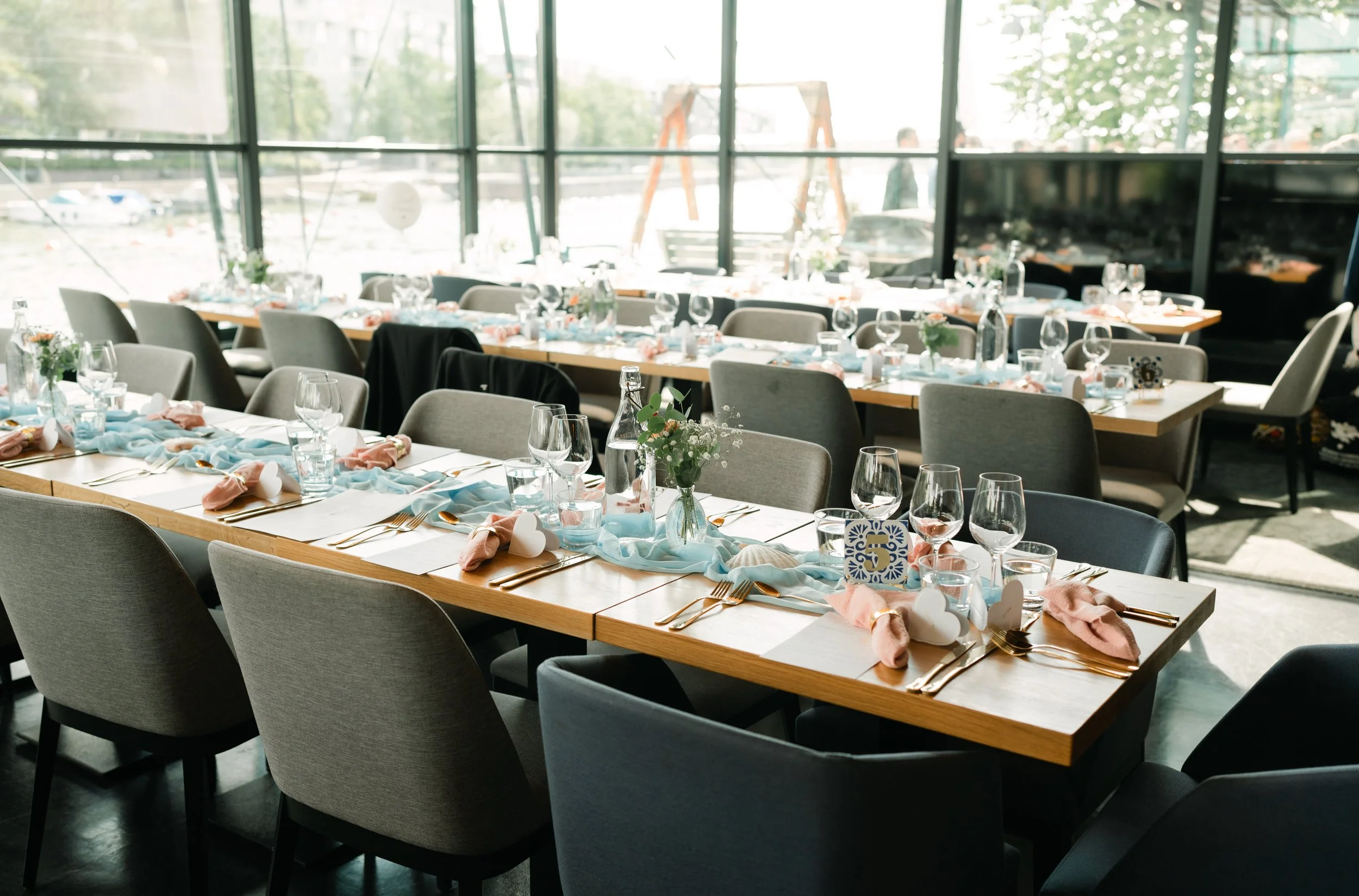 Elegant restaurant table setup with glasses, cutlery, napkins, and decorative elements in a sunlit dining area with large windows.