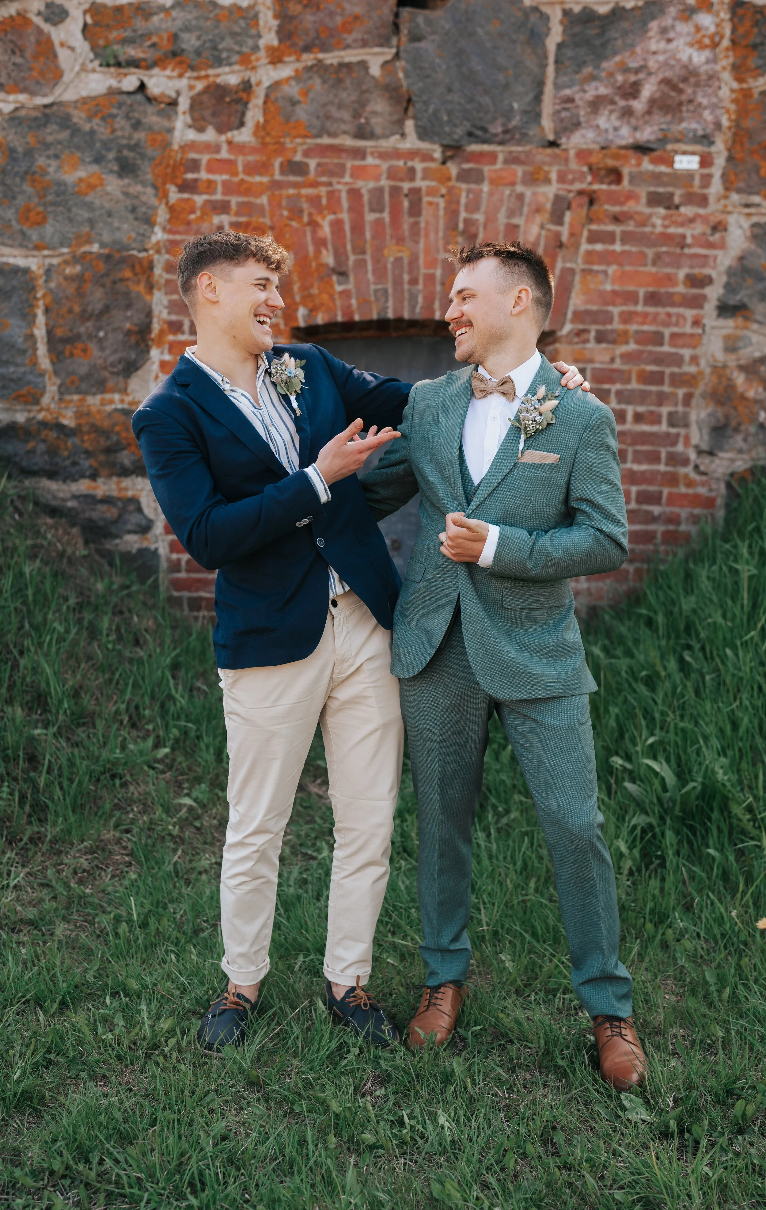 Two men in suits smiling and interacting in front of a brick wall.