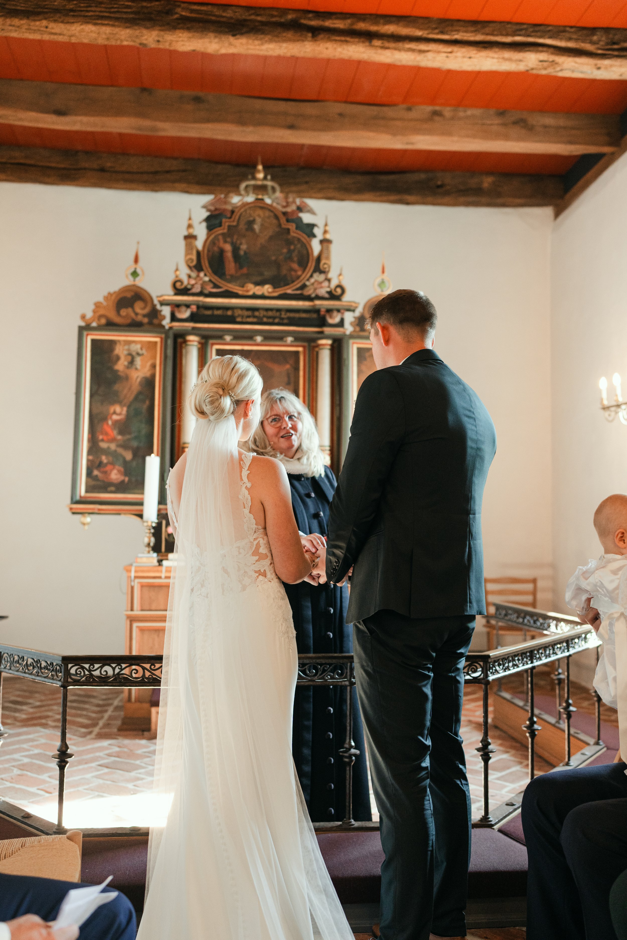 Bride and groom standing together during a wedding ceremony in a church, with an officiant in front of an ornate altar.