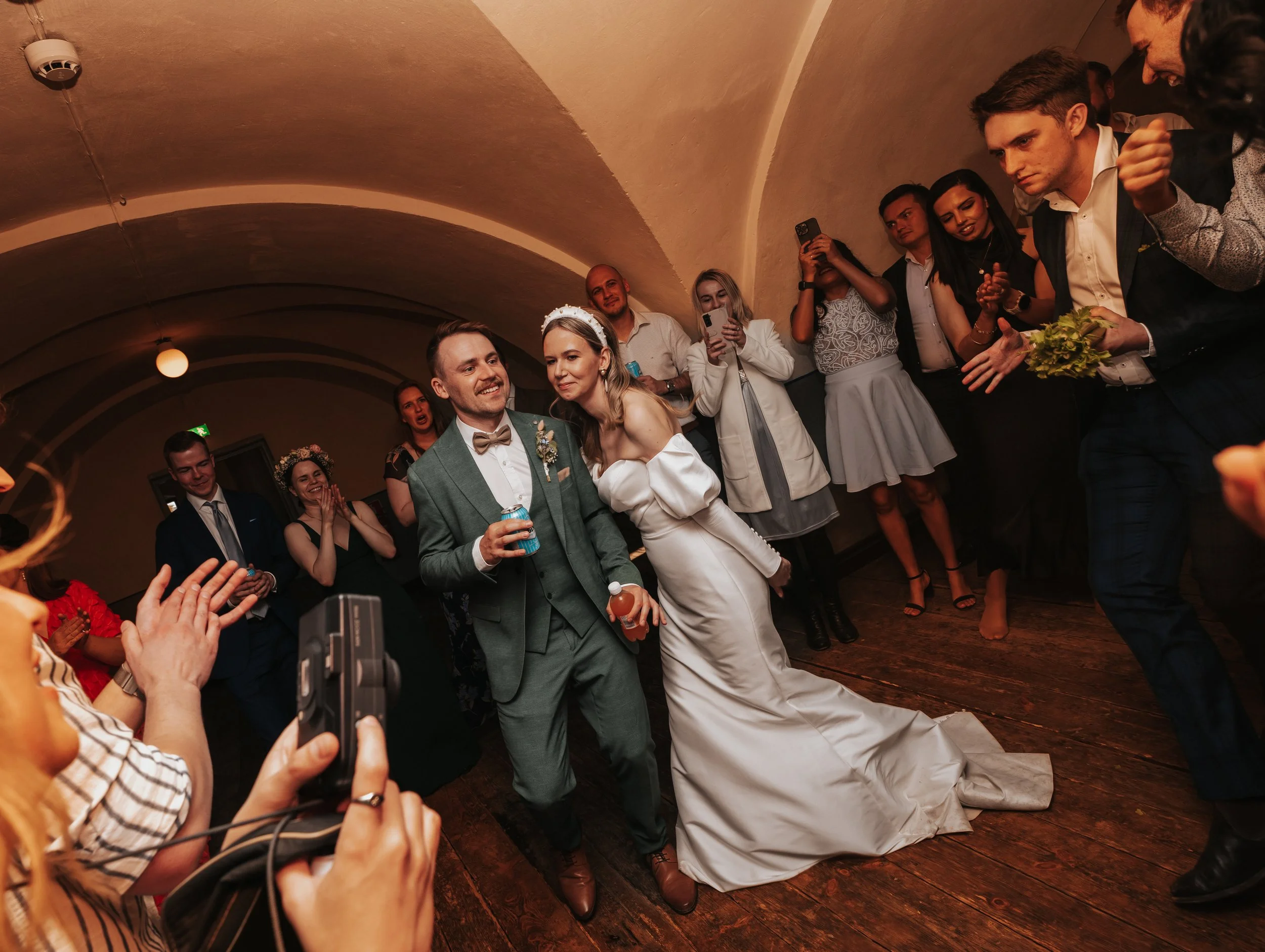 Bride and groom dancing with guests clapping at wedding reception, indoor celebration.