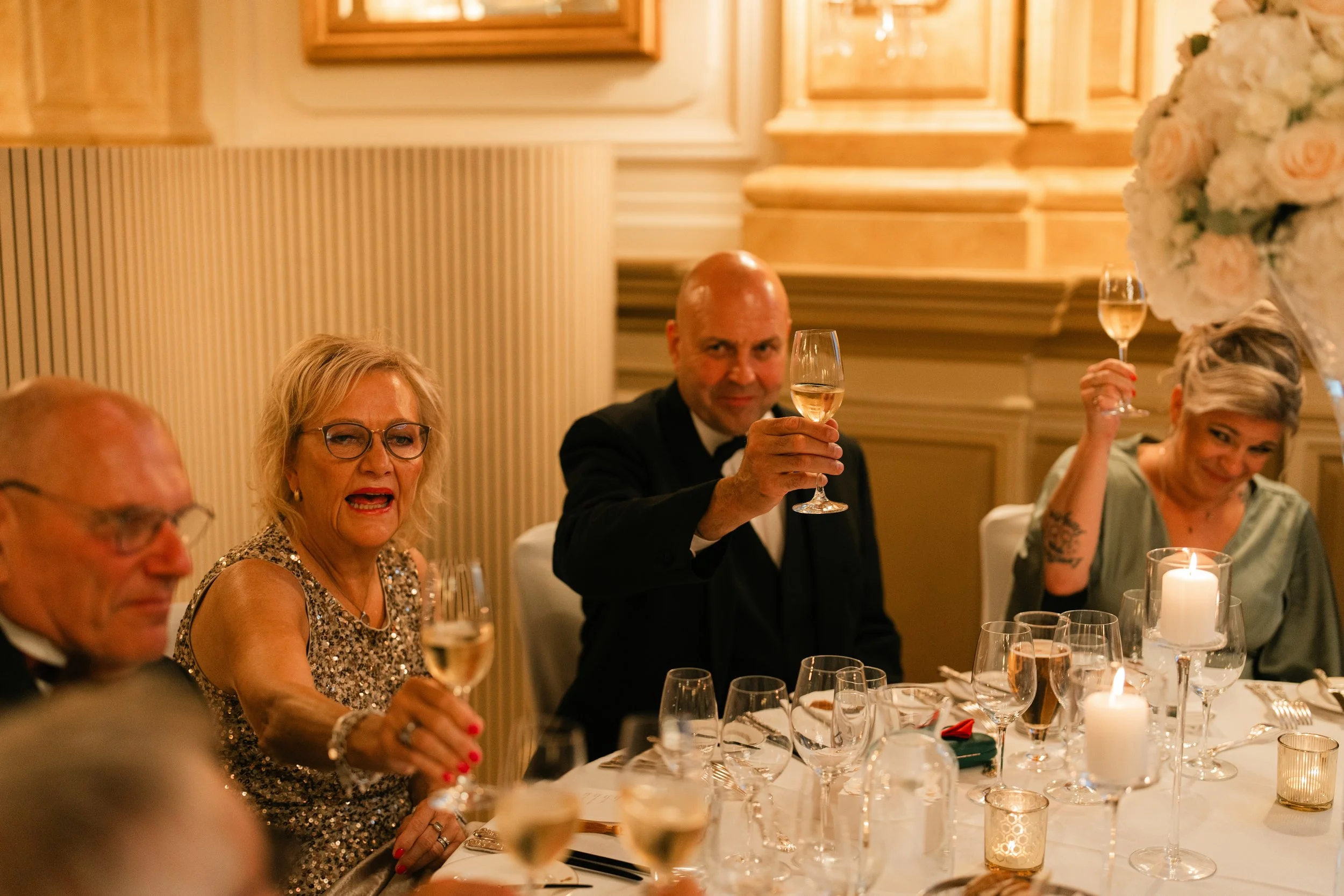 People raising glasses for a toast at an elegantly set dinner table with candles and flower arrangements.