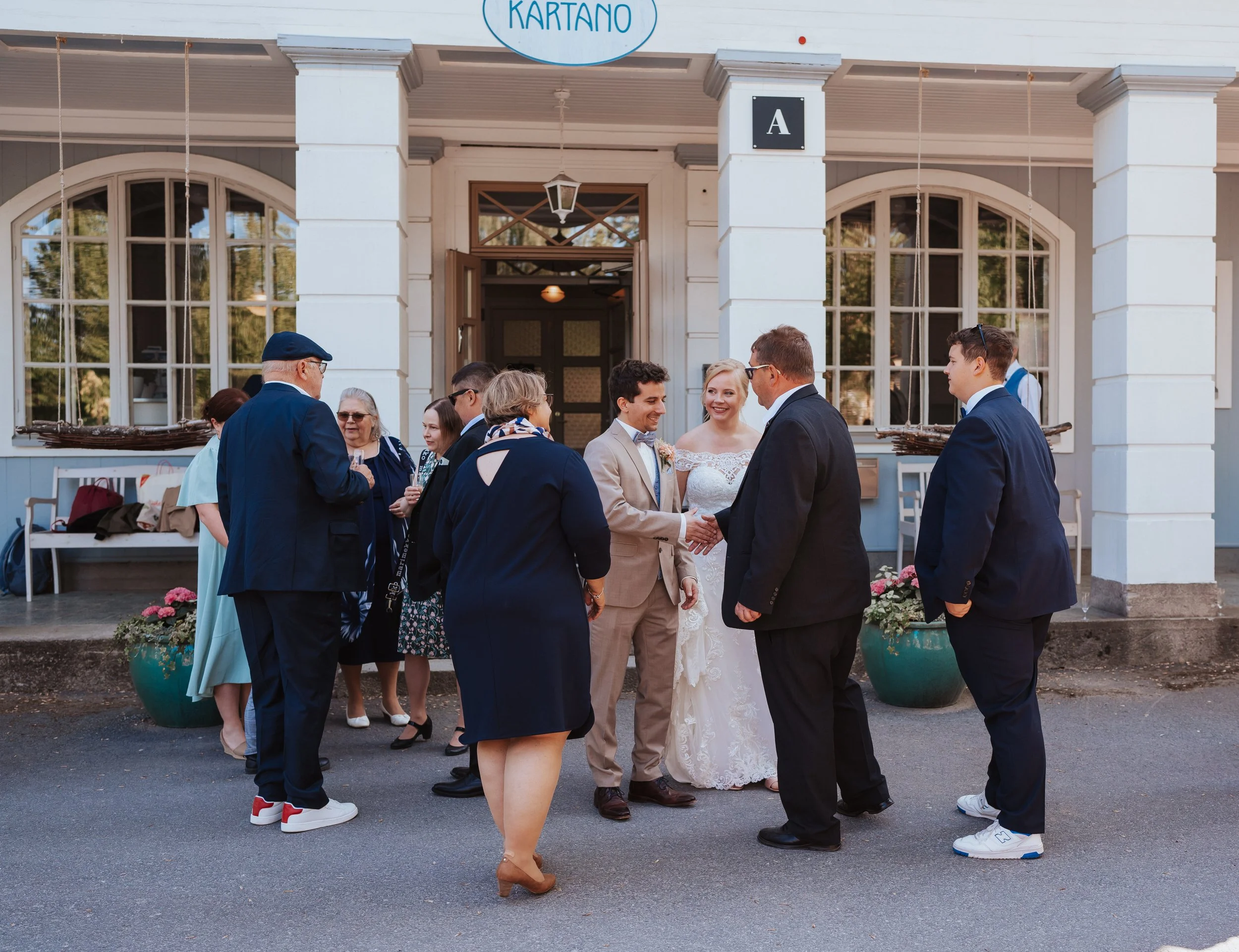 Group of people, including a bride and groom in wedding attire, conversing outside a building with arched windows and a sign labeled 'KARTANO'. Other guests are dressed in formal and business attire. Two large planters with flowers are visible. The s
