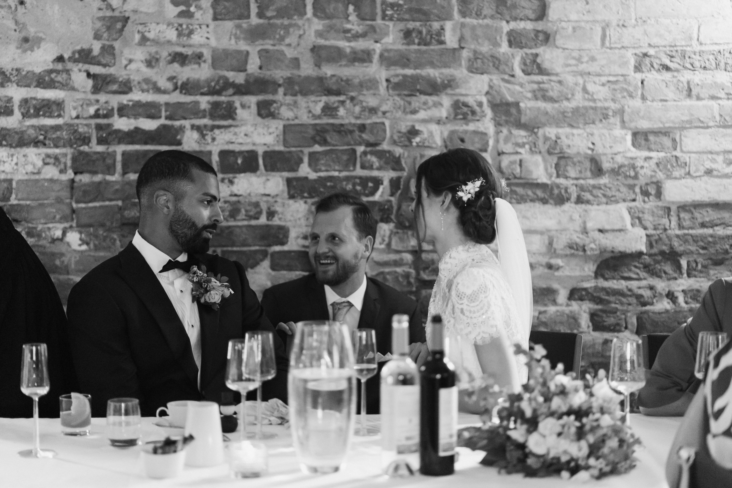 A black and white photo of three people in formal attire sitting at a table in front of a brick wall. They appear to be having a conversation during a wedding reception. The table is set with glasses and bottles, and there is a floral arrangement.