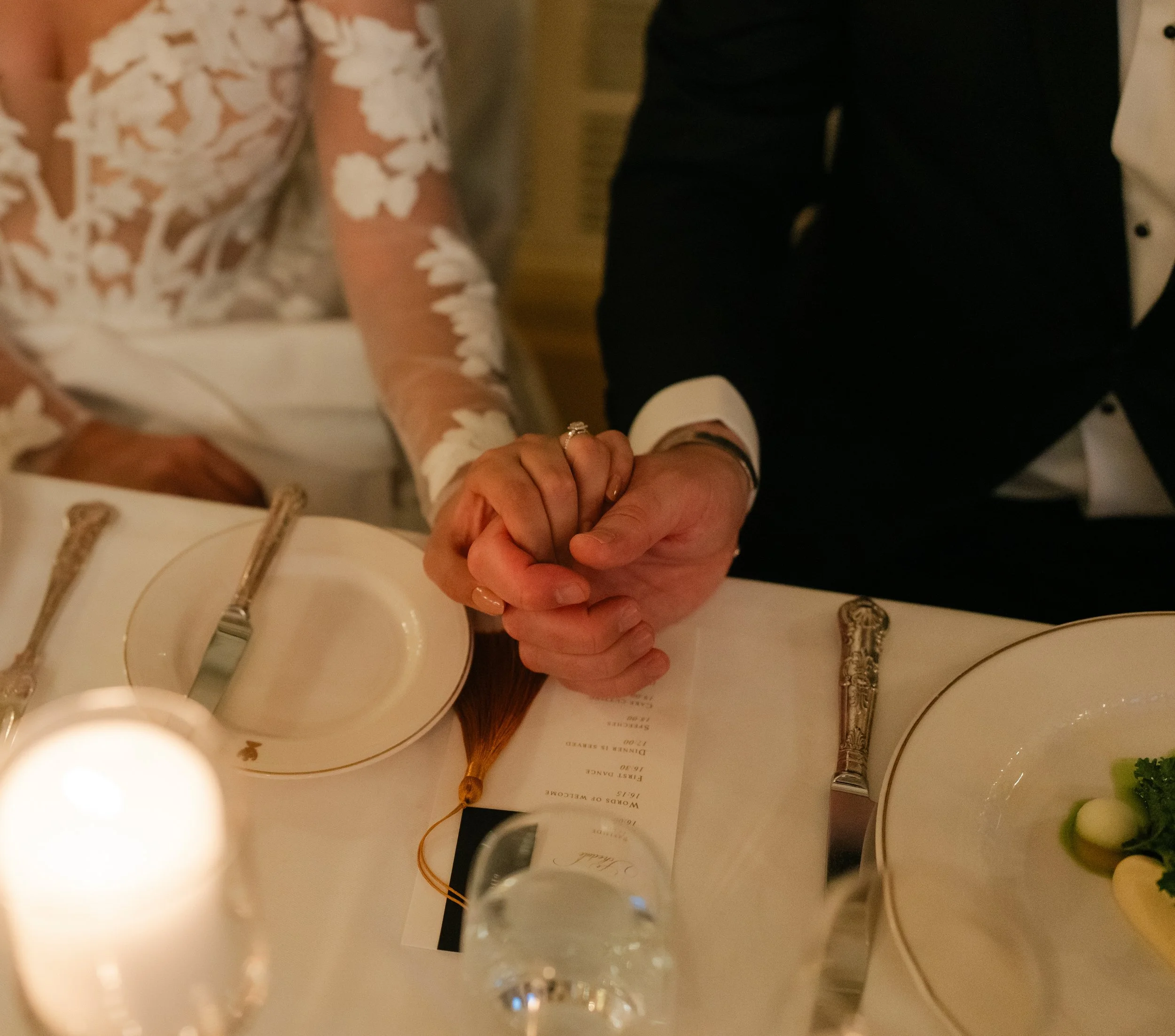 Bride and groom holding hands at a wedding reception table with elegant place settings and a candle.