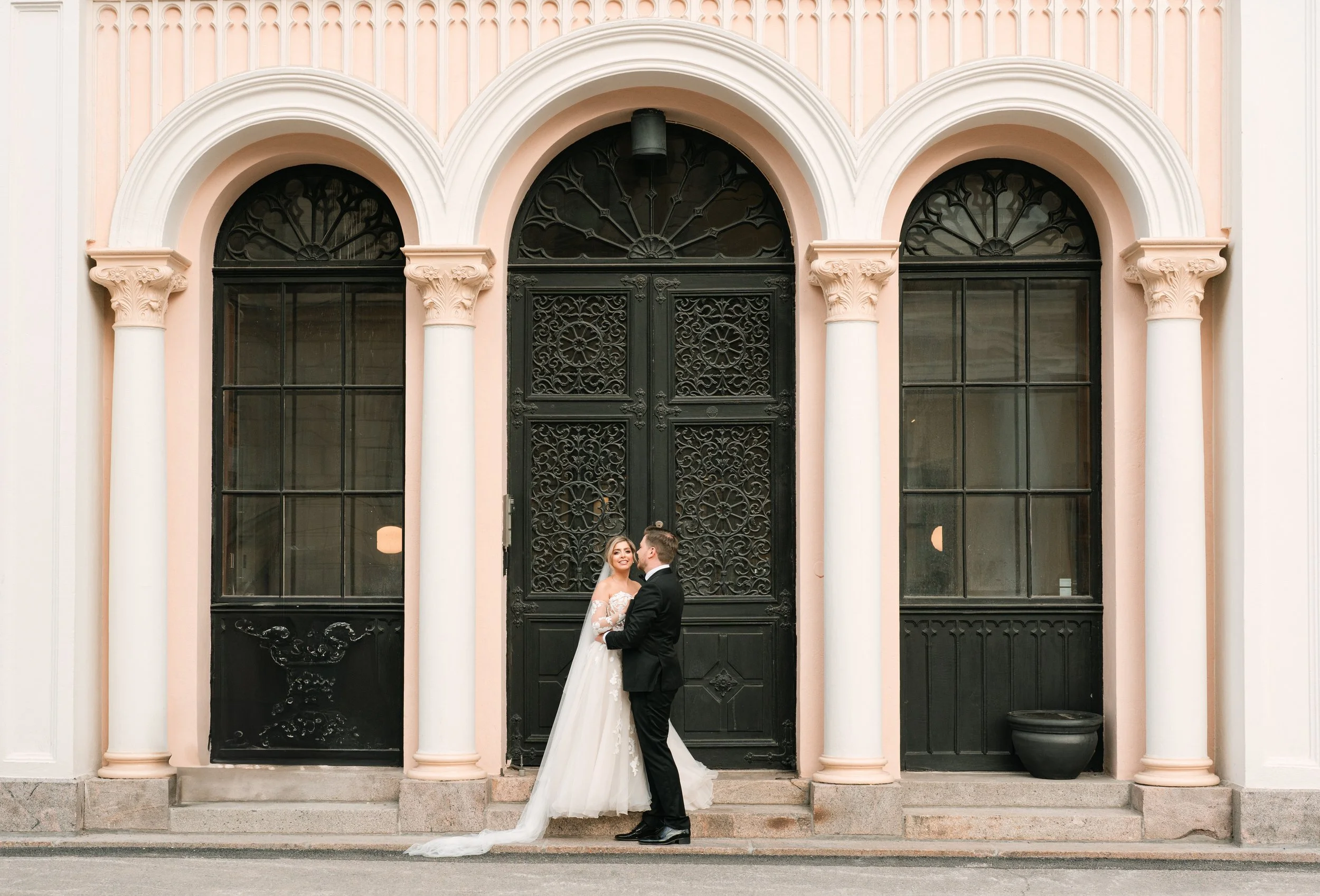 Bride and groom embracing in front of ornate building with arched windows and columns.